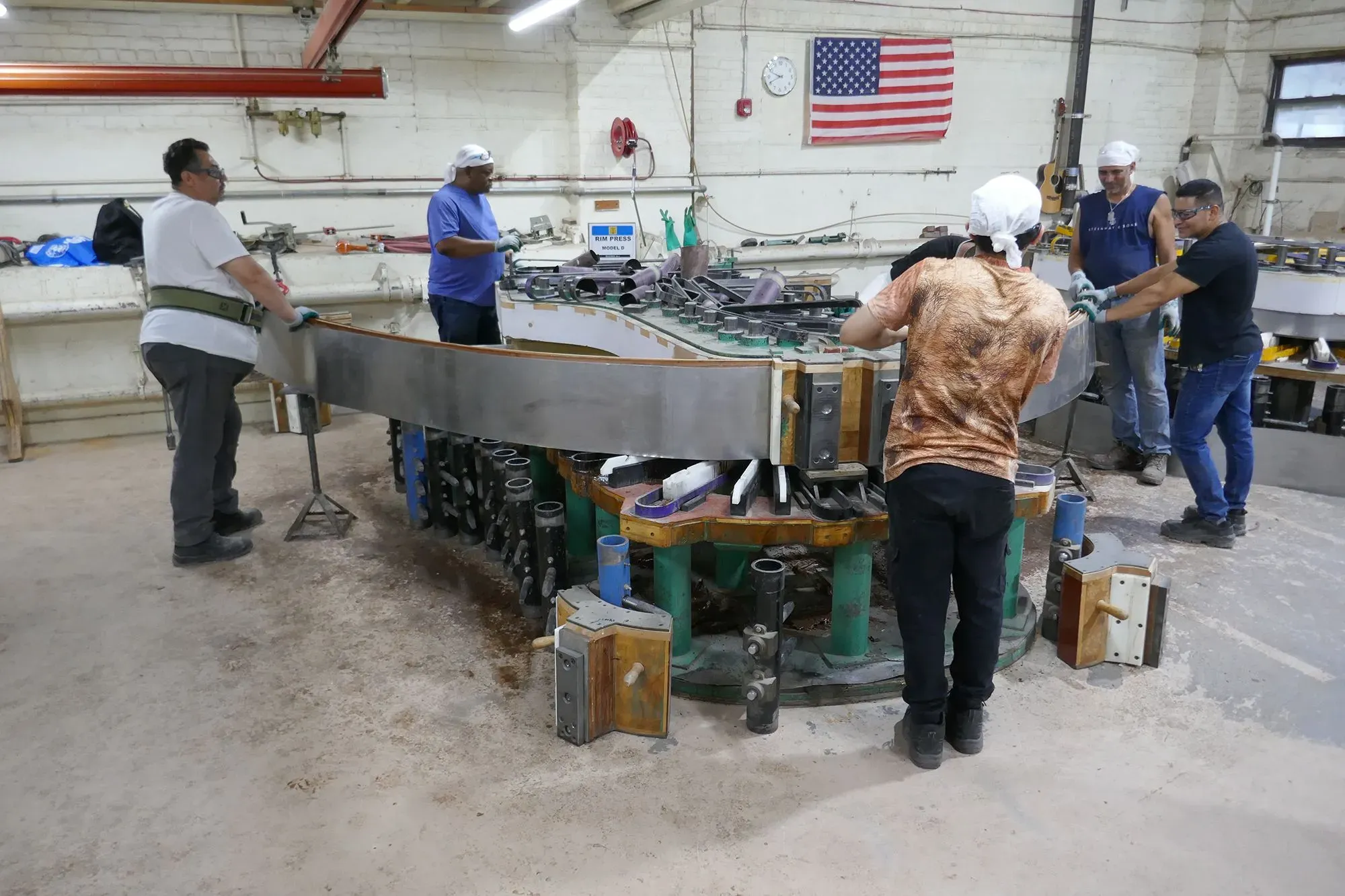 عمال في مصنع شتاينواي وأولاده يقومون بتشكيل إطار بيانو باستخدام أدوات متنوعة، مع العلم الأمريكي في الخلفية، مما يعكس الحرفية العالية في صناعة البيانو.