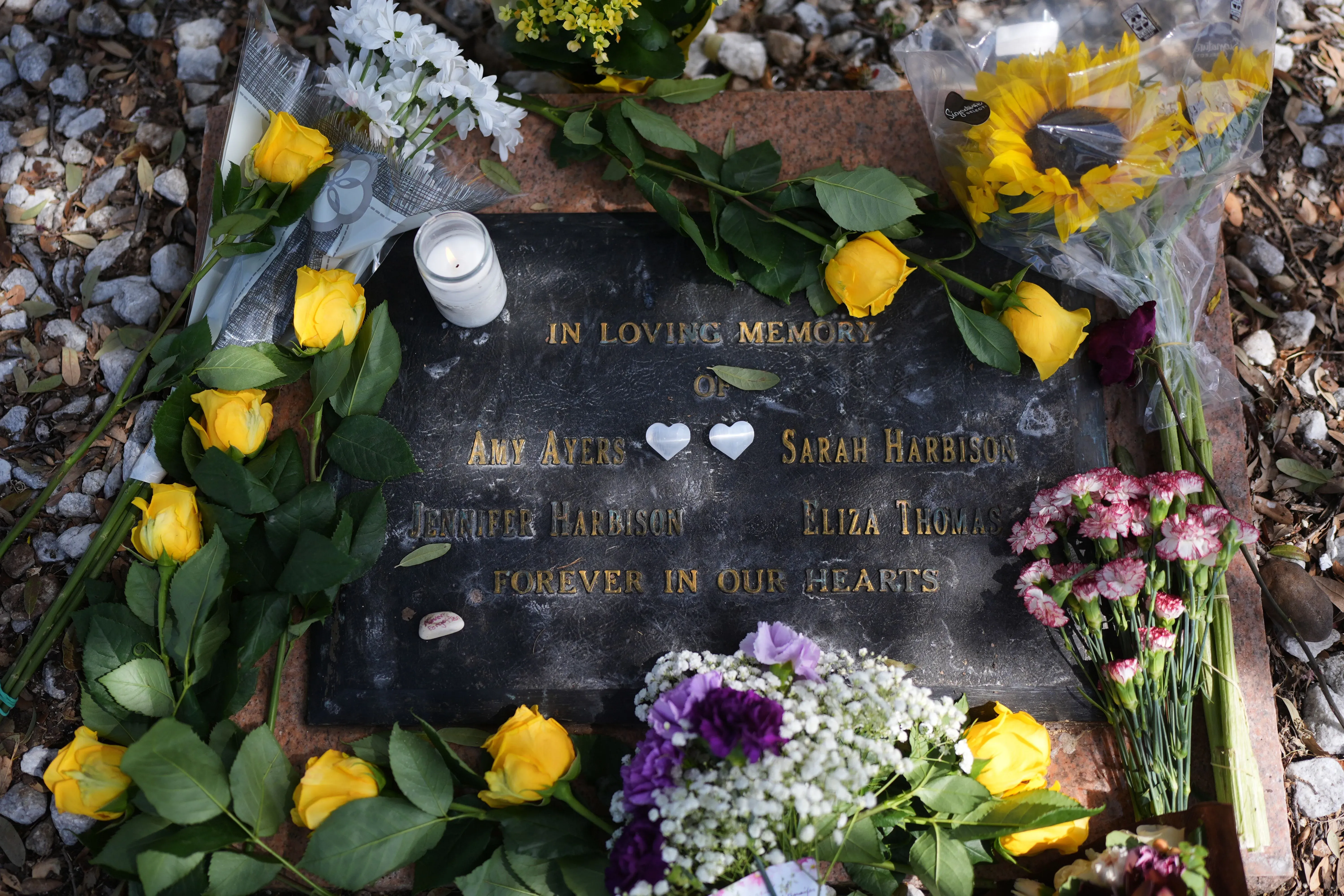 شاهد لافتة تذكارية تحمل أسماء أربع فتيات مراهقات تم قتلهن في عام 1991، محاطة بالورود والشموع، تعبيرًا عن الذكرى والاحترام.