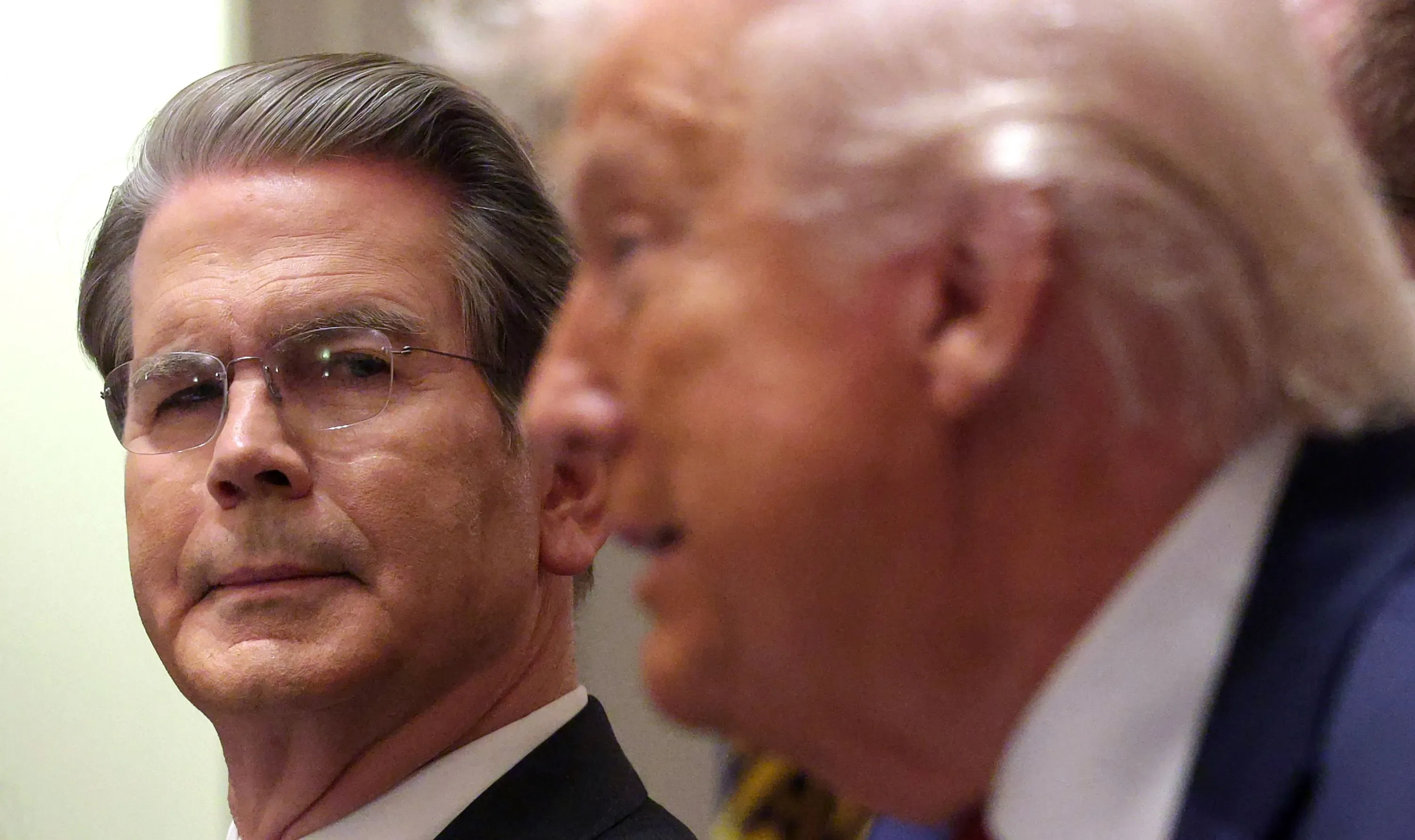 بيسنت يستمع إلى ترامب خلال مؤتمر صحفي، مع تعبير جاد يعكس دوره كمدافع عن السياسات الاقتصادية للإدارة الأمريكية.