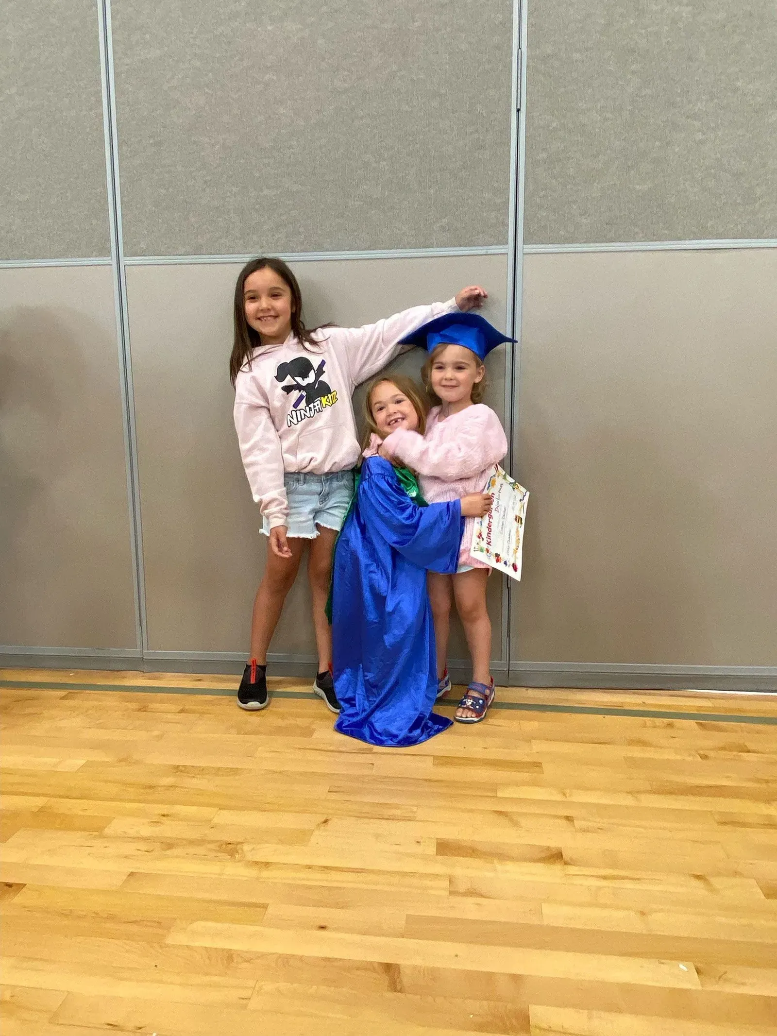 ثلاث فتيات صغيرات يتظاهرن بالاحتفال، إحداهن ترتدي قبعة تخرج وزي تخرج أزرق، بينما تبتسم الأخريات في خلفية بسيطة.