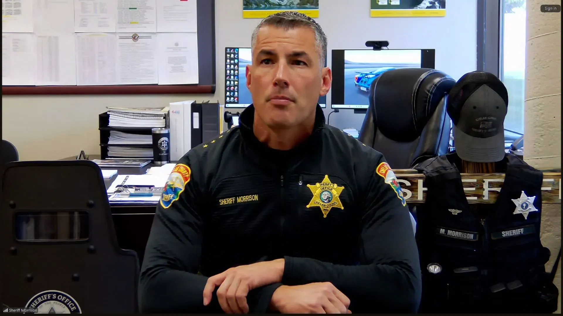 محقق يجلس في مكتبه، يرتدي زي الشرطة، مع خلفية تحتوي على مستندات وأغراض مكتبية، يتحدث عن قضية ترافيس ديكر.