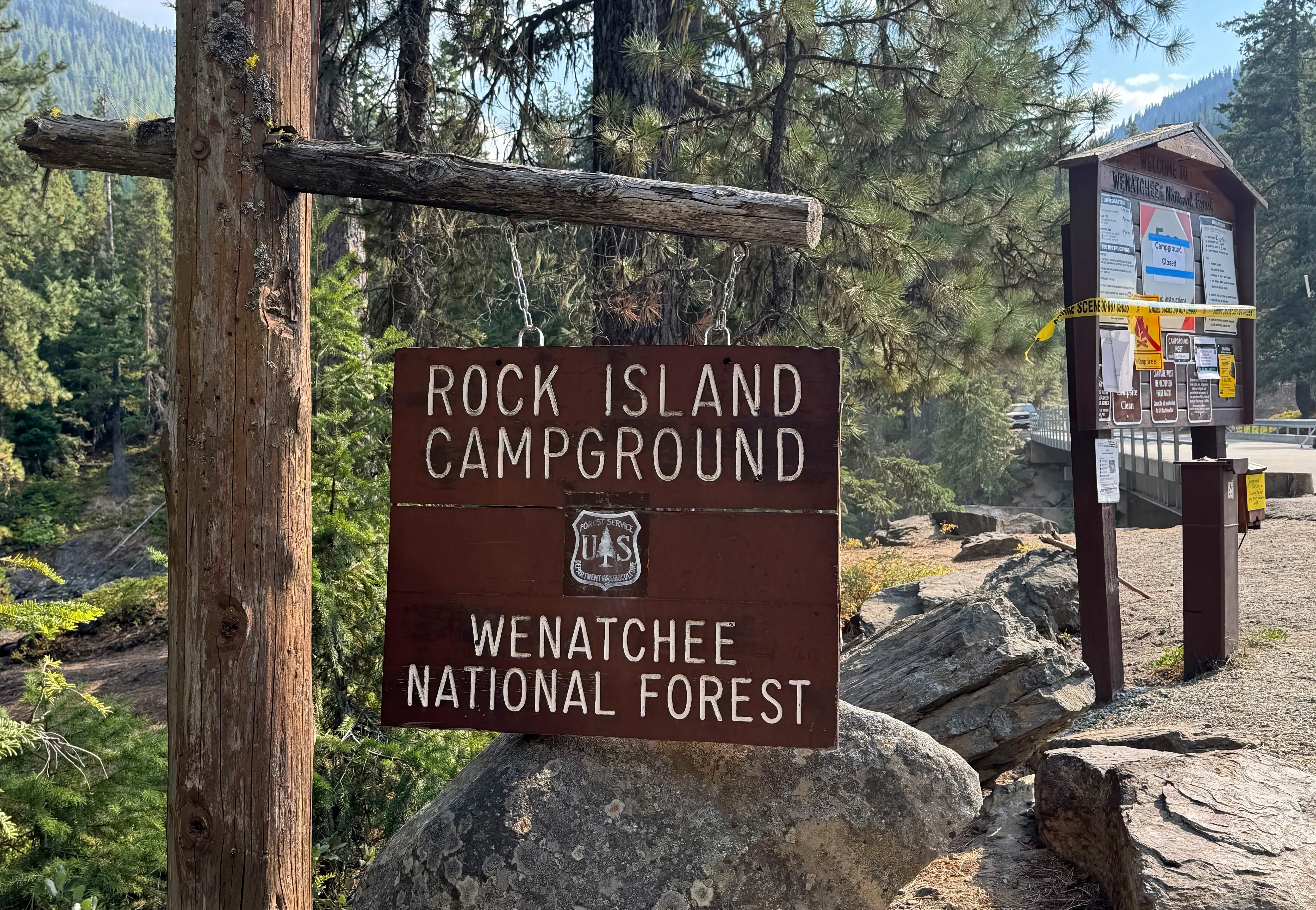 علامة تشير إلى مخيم روك آيلاند في غابة ويناتشي الوطنية، محاطة بأشجار كثيفة، مع وجود شريط تحذيري يشير إلى وجود تحقيقات جارية.