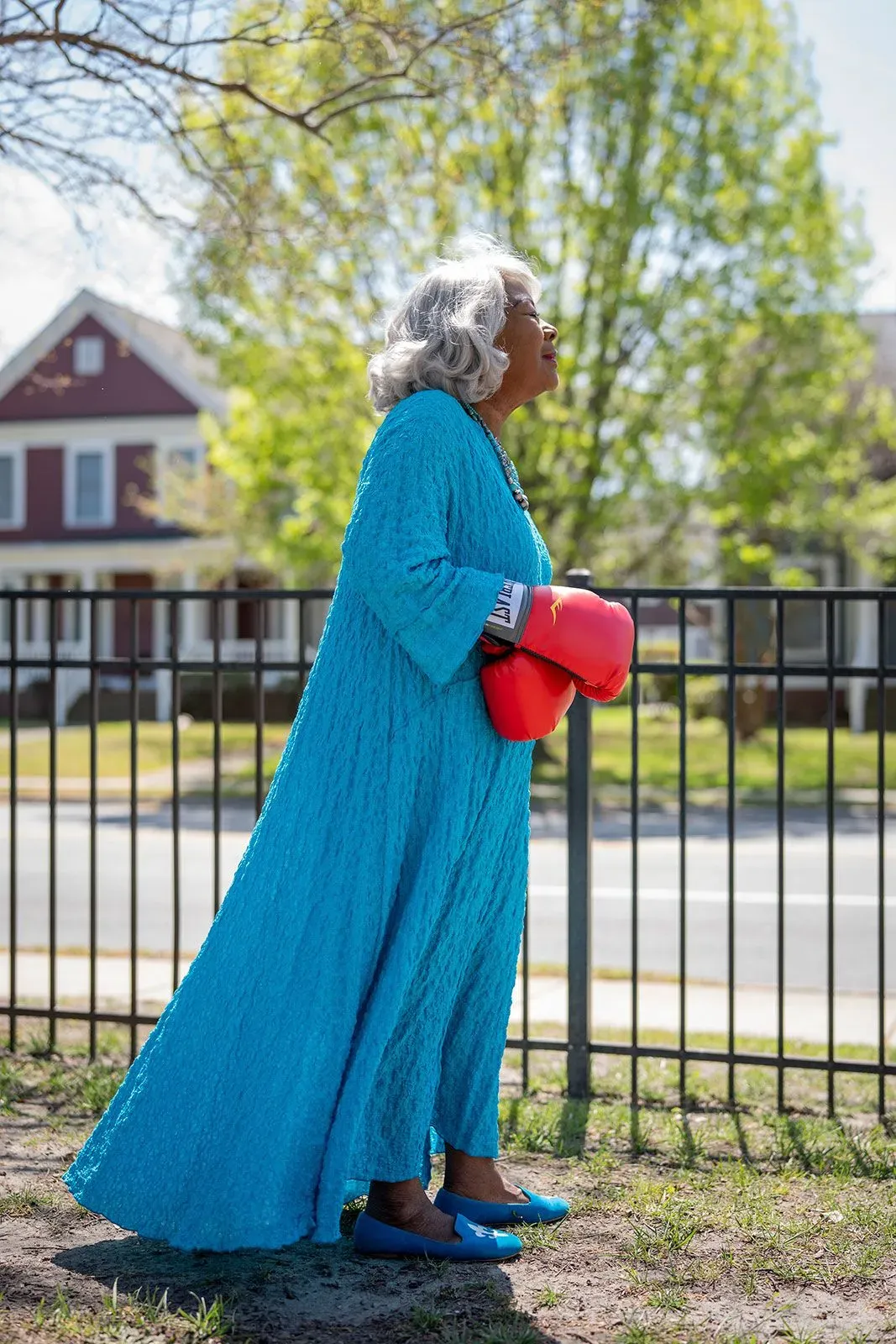 امرأة مسنّة ترتدي فستانًا أزرق طويلًا وتحمل قفازات ملاكمة حمراء، تتجه نحو الكاميرا في مشهد يعكس قوتها السياسية وعزمها في مواجهة التحديات.