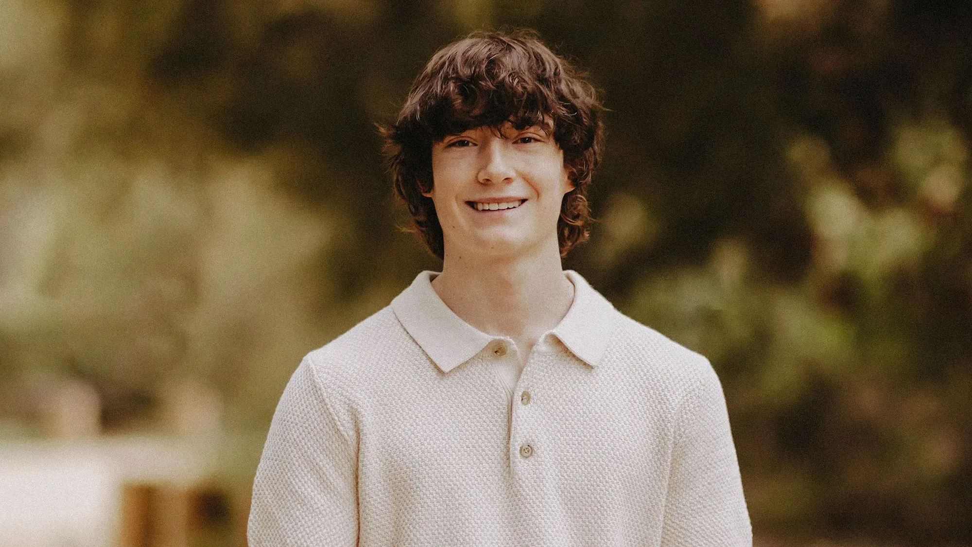 شاب مبتسم يرتدي قميصًا بأزرار، يقف في بيئة طبيعية، مما يعكس موضوع الصحة العقلية وتأثير الذكاء الاصطناعي على المراهقين.