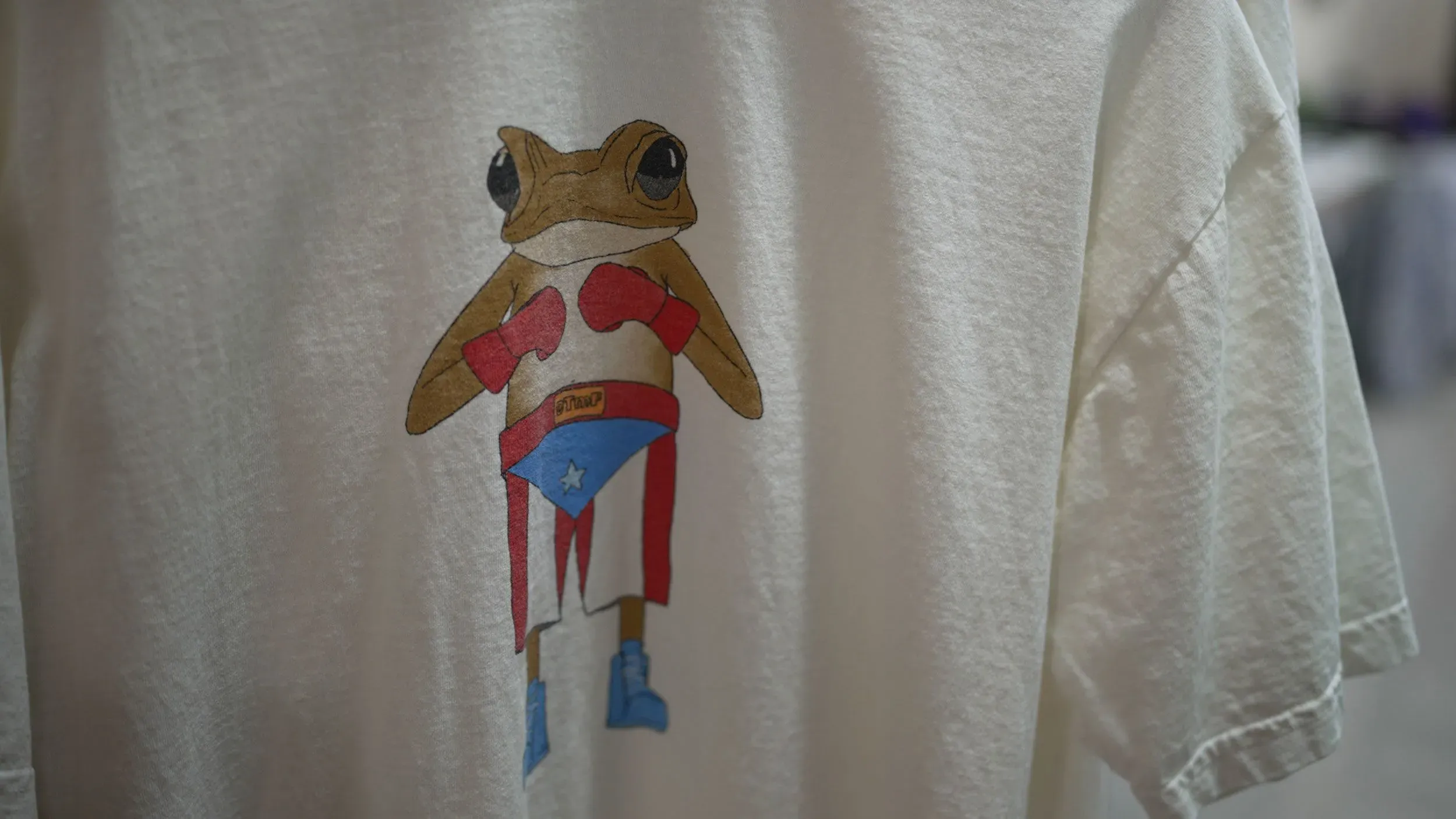 قميص أبيض يحمل رسمة ضفدع يرتدي قفازات ملاكمة ويرمز إلى الثقافة البورتورية، في سياق حفلات باد باني في بورتوريكو.