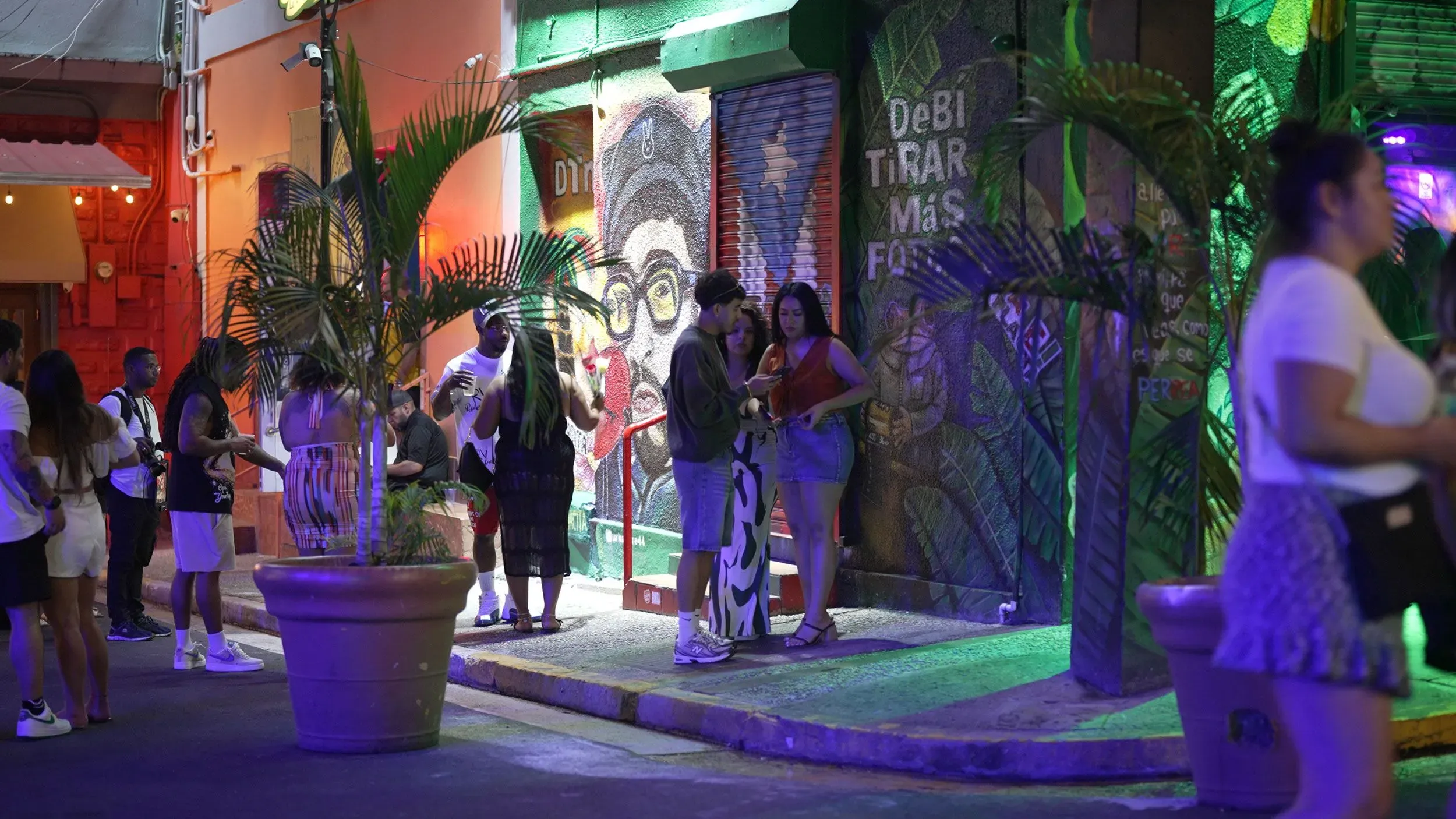 مجموعة من الأشخاص يقفون في شارع ملون في سان خوان، بورتوريكو، بالقرب من جدارية لباد باني، مع أجواء احتفالية تعكس تأثيره الثقافي.