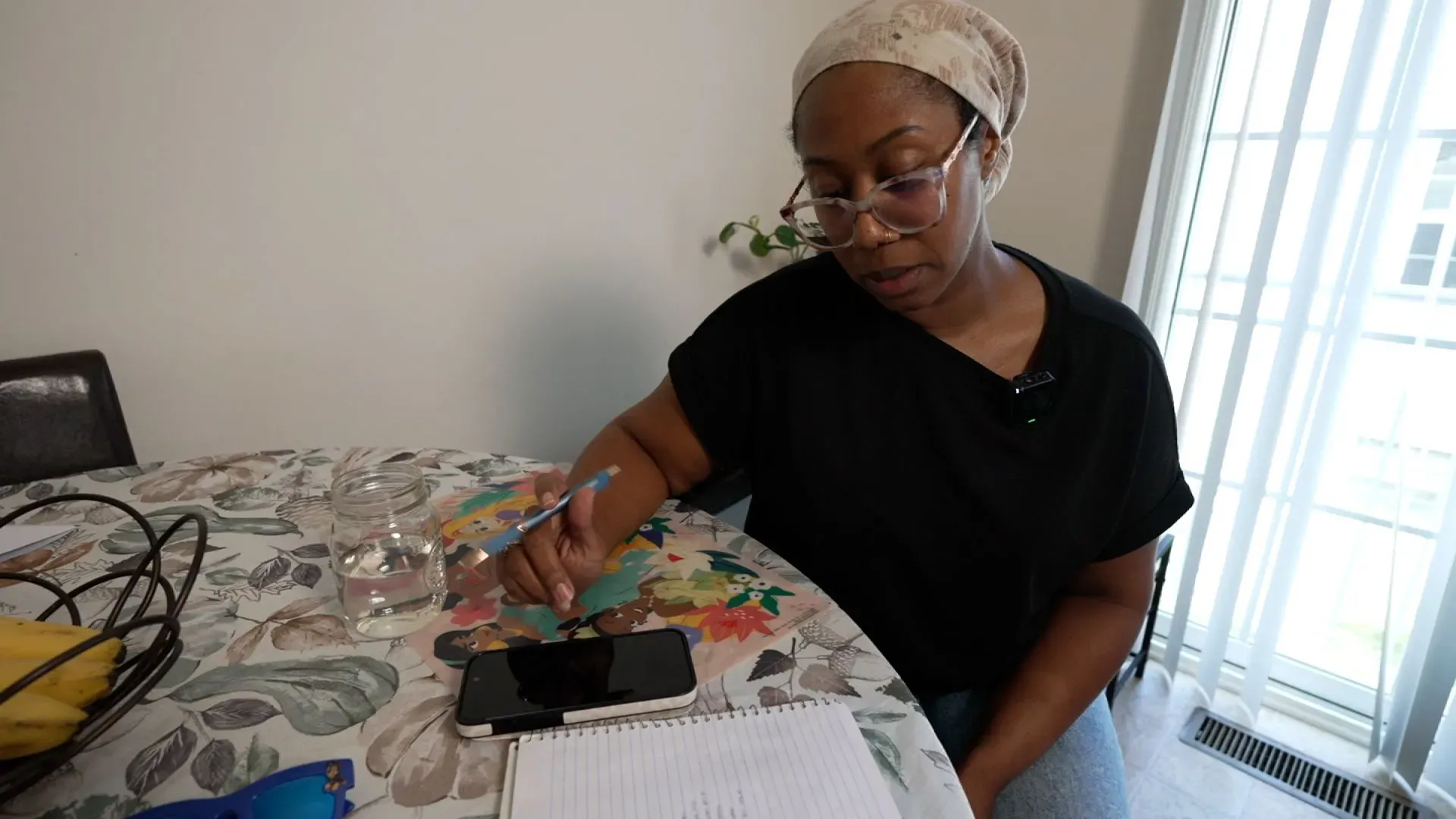 امرأة ترتدي نظارات وتكتب على دفتر ملاحظات بجانب هاتفها، تعبر عن القلق بشأن ميزانيتها الغذائية. تعكس الصورة التحديات التي تواجهها الأسر بسبب نقص الدعم من برنامج SNAP.