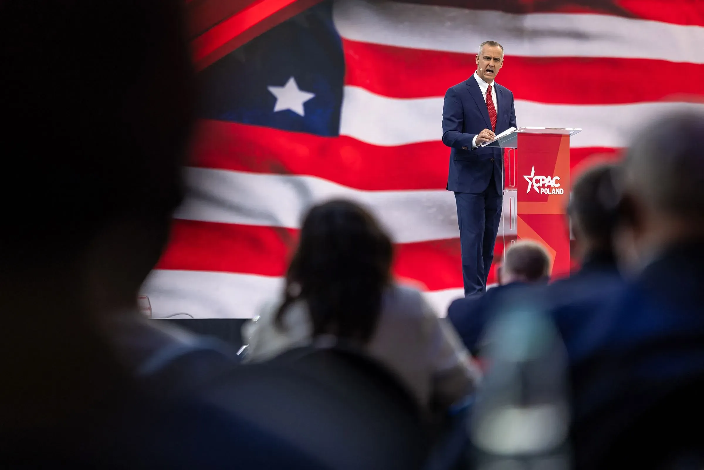 كوري ليفاندوفسكي يتحدث في مؤتمر مع خلفية علم أمريكي، مشيرًا إلى نفوذه في وزارة الأمن الداخلي خلال جولة وزيرة الأمن الداخلي في أمريكا الجنوبية.