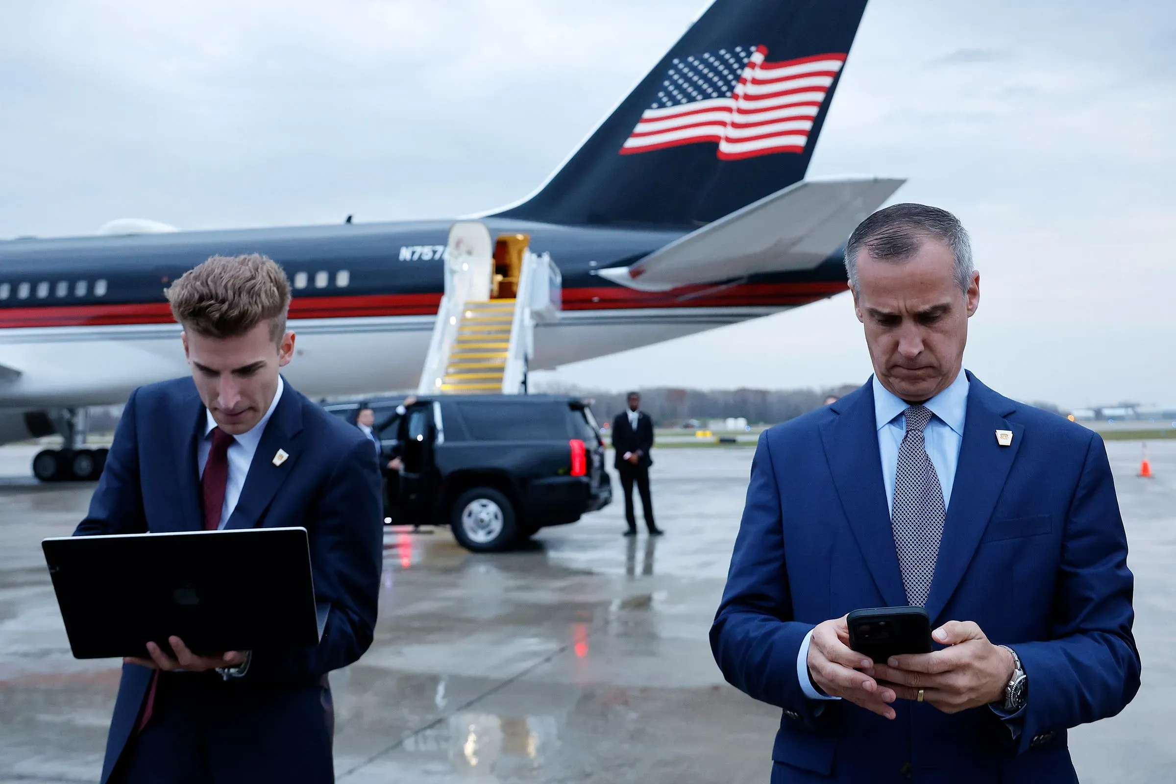 كوري ليفاندوفسكي يتفقد هاتفه بينما يعمل مساعده على اللابتوب، خلفهما طائرة تحمل علم الولايات المتحدة، خلال رحلة إلى أمريكا الجنوبية.