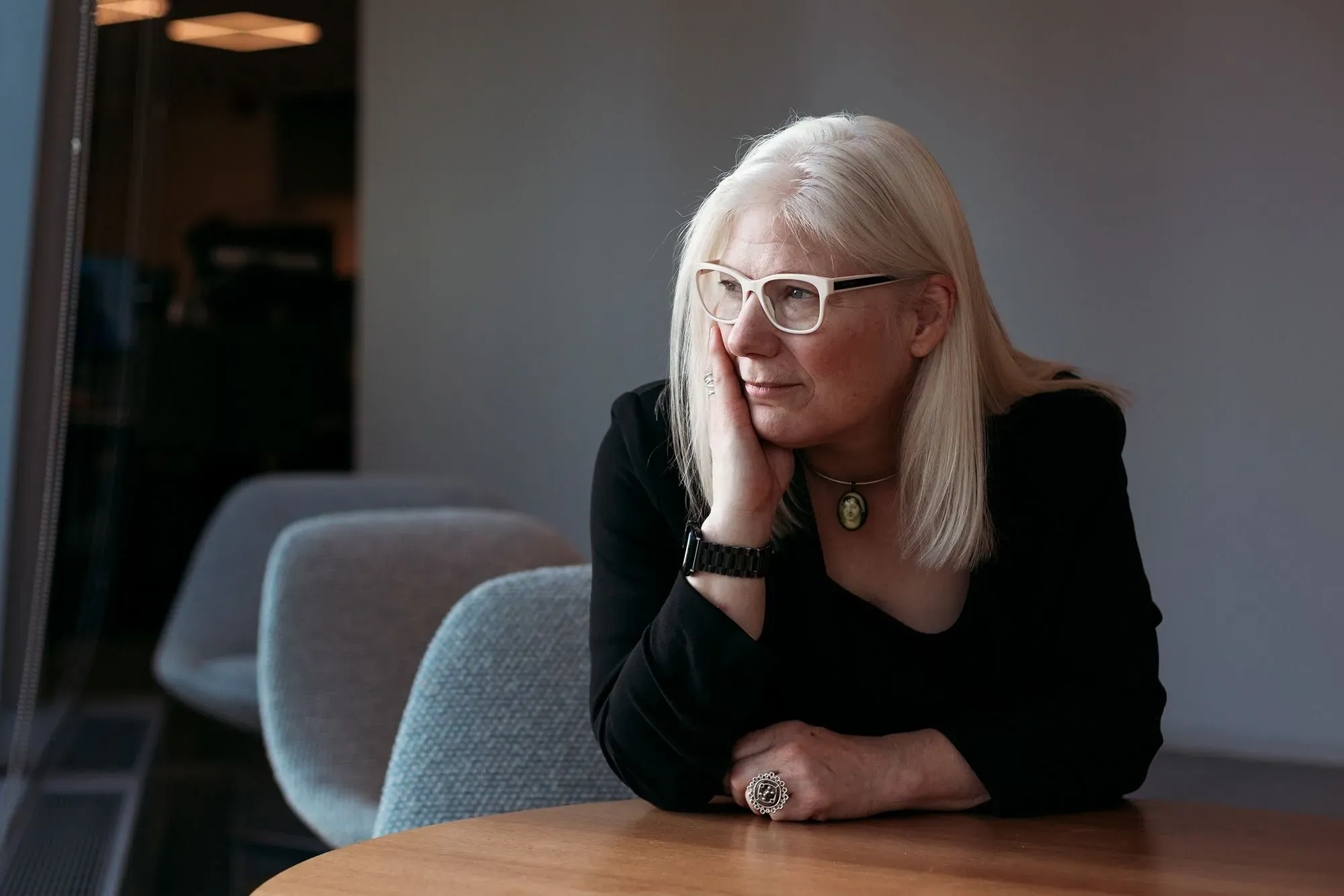 امرأة ذات شعر أشقر طويل ترتدي نظارات، تجلس على طاولة وتفكر بعمق. تعكس تعابير وجهها اهتمامًا بقصة جين أوستن وأيامها الأخيرة.