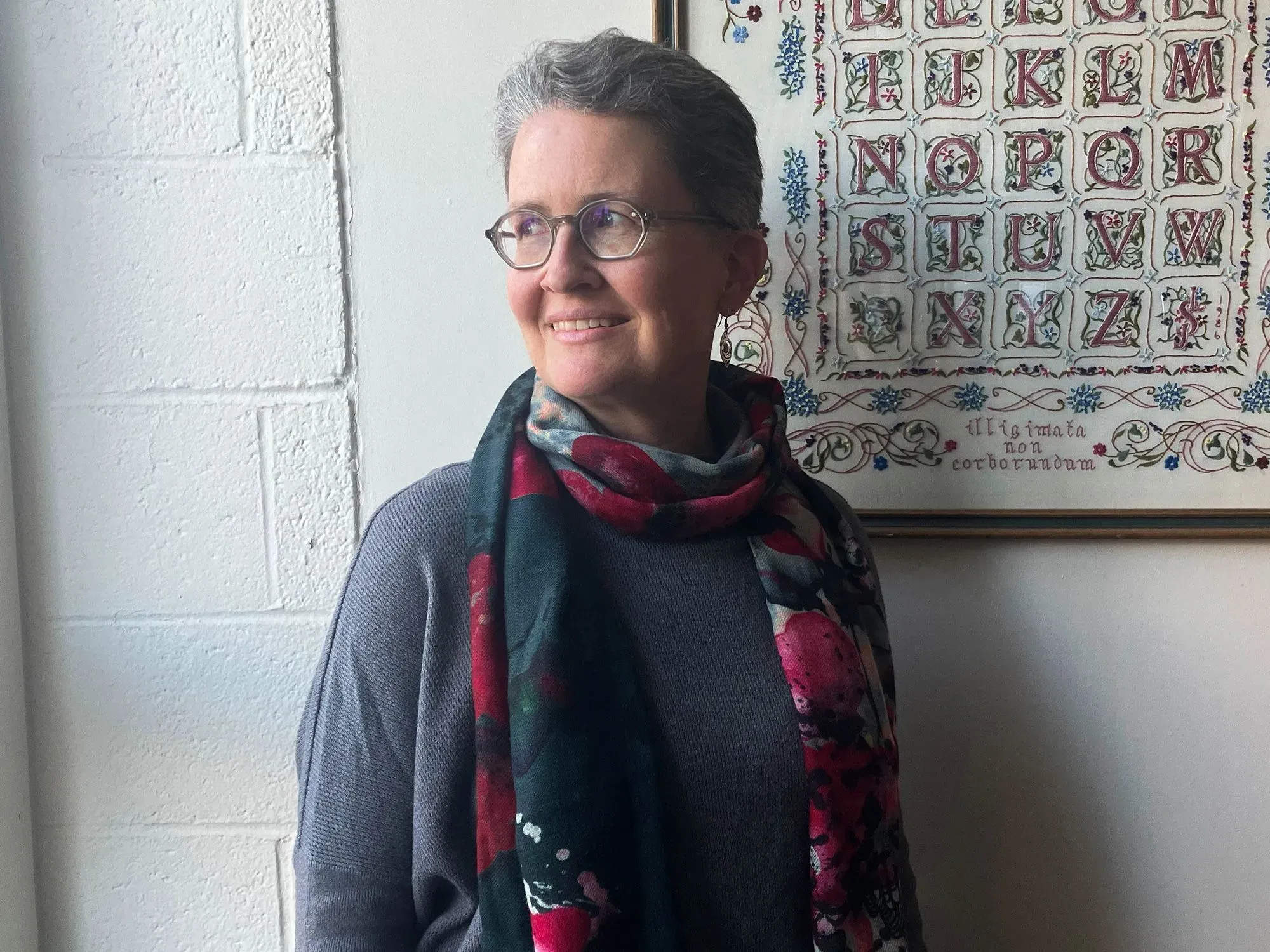 امرأة ترتدي سترة رمادية وشال مزخرف، مبتسمة أمام جدار أبيض مزين بإطار يحتوي على حروف مزخرفة.