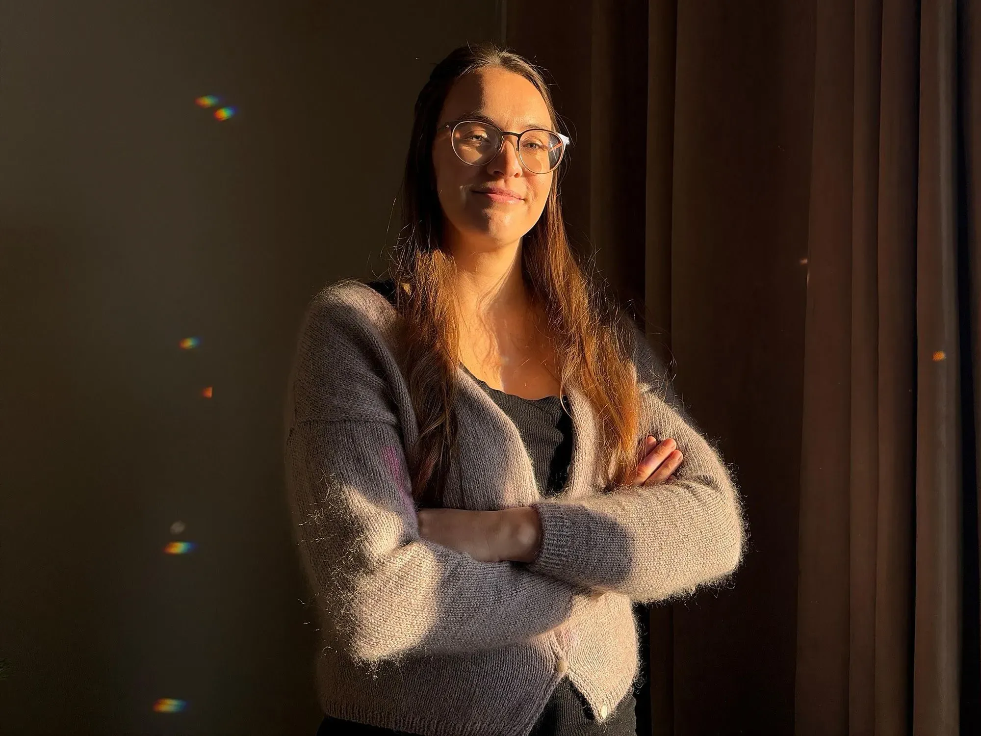 امرأة مبتسمة ترتدي نظارات وقميص أسود مع سترة رمادية، تقف أمام نافذة تسمح بدخول الضوء، مما يخلق تأثيرات لونية على الجدار.