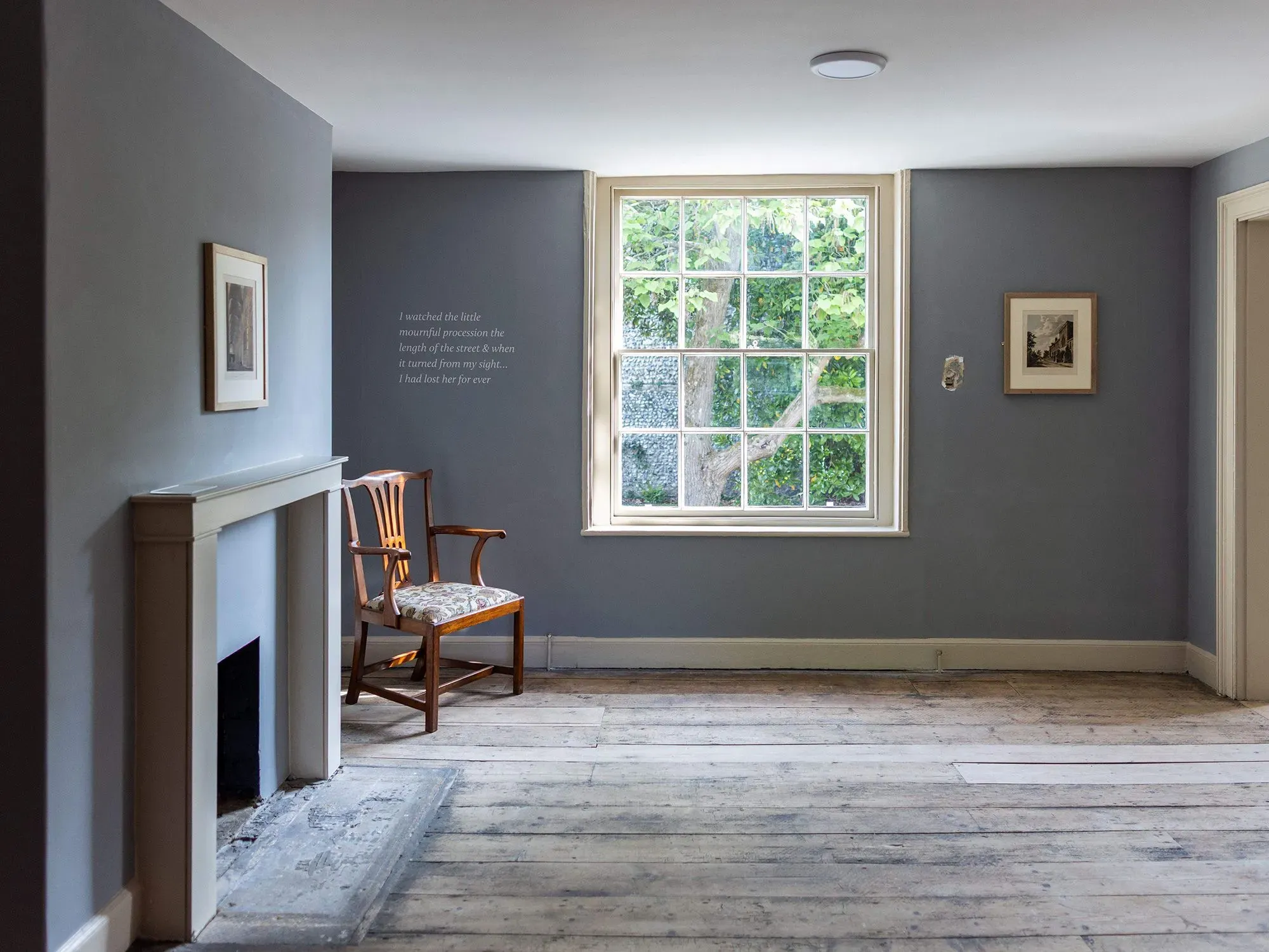 غرفة بسيطة مع جدار رمادي ونافذة كبيرة، تحتوي على كرسي خشبي ومقتنيات فنية، تعكس حياة جين أوستن في آخر أيامها.