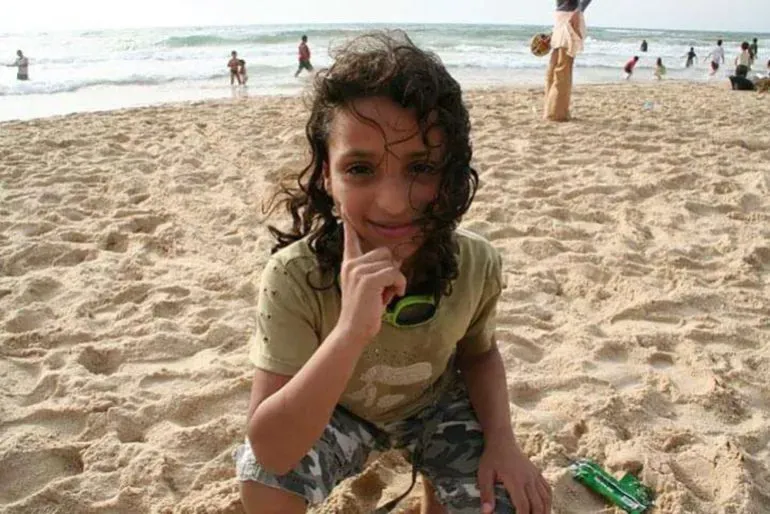 طفلة صغيرة على شاطئ غزة، ترتدي قميصًا أخضر وتبتسم، بينما تلعب مع عائلتها في الرمال بالقرب من البحر.