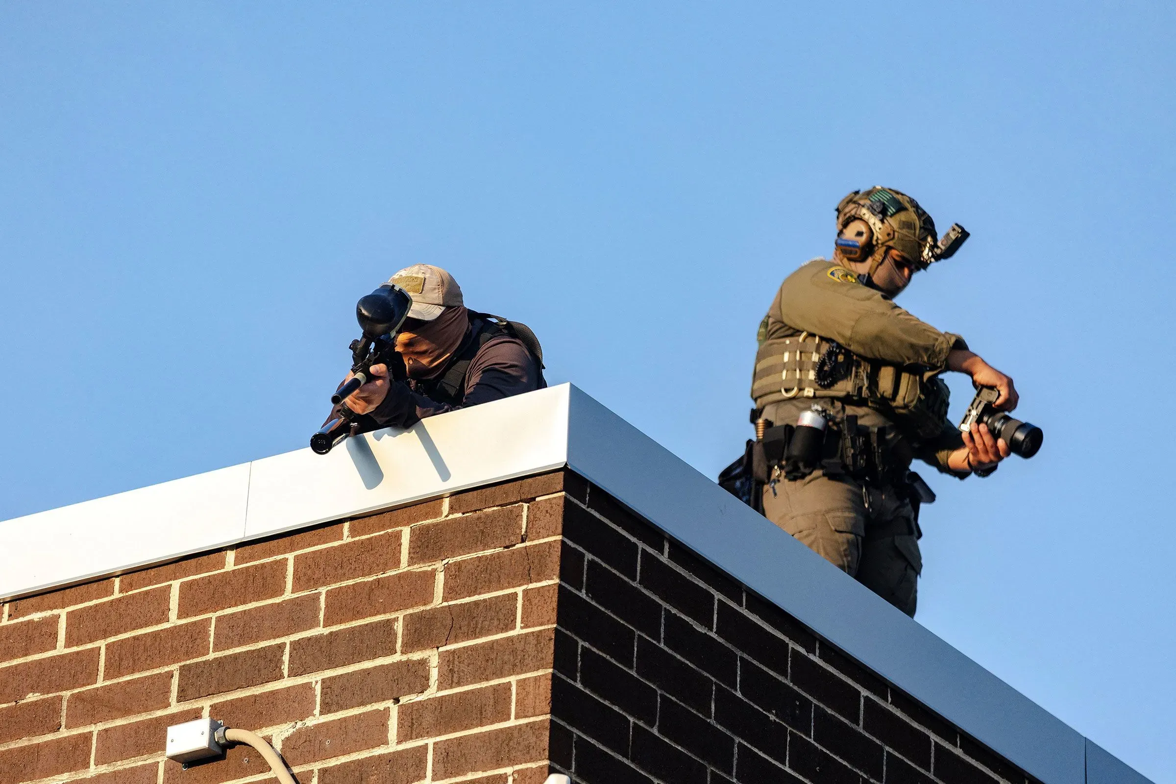 عملاء فيدراليون مسلحون بأسلحة أقل فتكًا، يراقبون من سطح مبنى خلال مظاهرة، مما يعكس تصاعد التوترات بين المتظاهرين والسلطات.