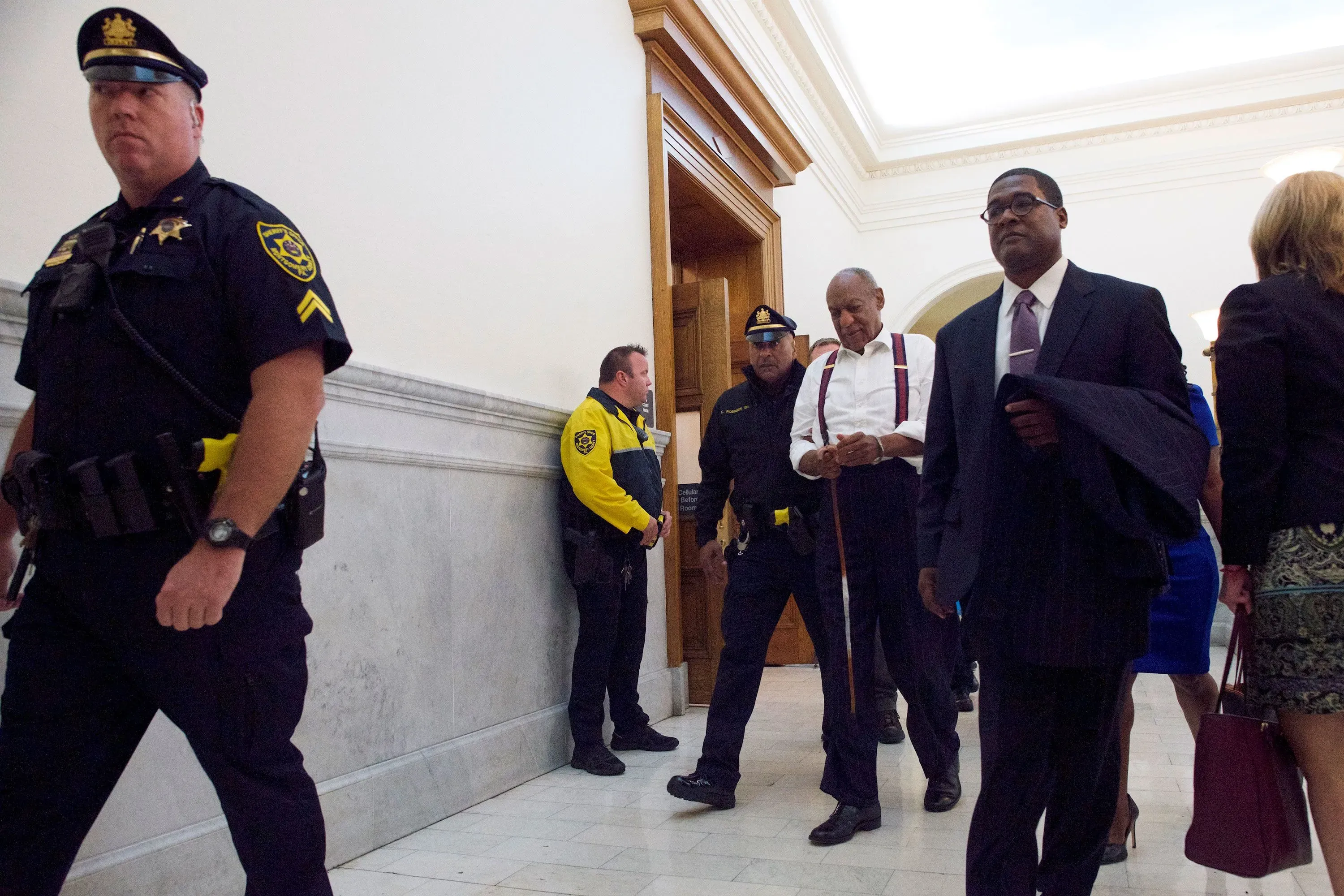 صورة لمجموعة من رجال الشرطة في قاعة المحكمة، حيث يظهر أحدهم يرتدي زيًا رسميًا، بينما يتجه شخص آخر نحو المدخل، مما يعكس أجواء المحاكمة المتعلقة بقضايا الاعتداء الجنسي.