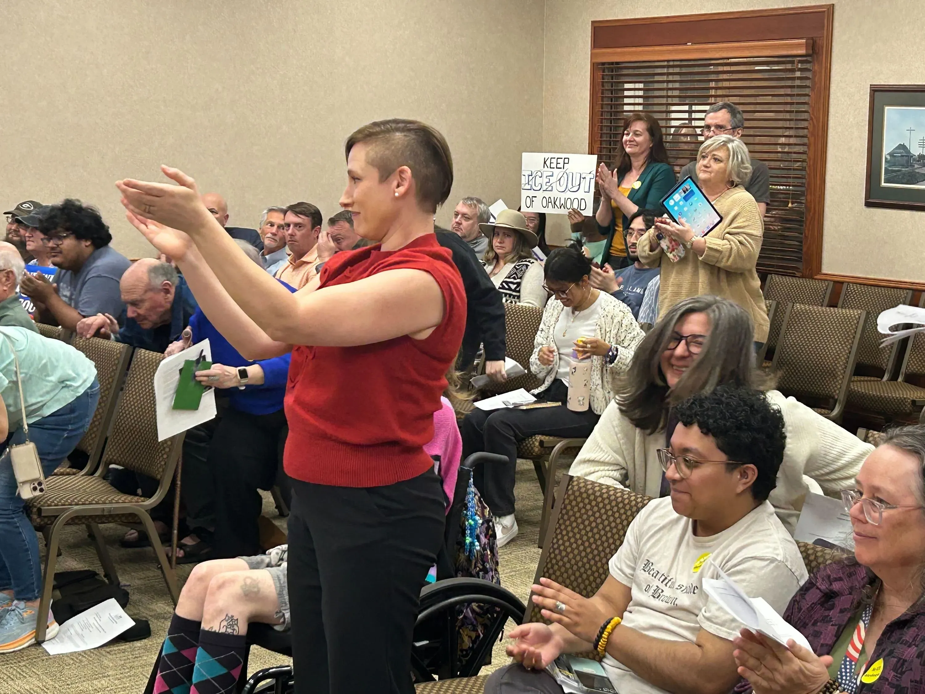 مجموعة من الأشخاص في اجتماع عام، يتفاعلون مع المتحدثة التي ترتدي سترة حمراء، بينما يحمل البعض لافتات تعبر عن معارضتهم لخطط احتجاز المهاجرين.