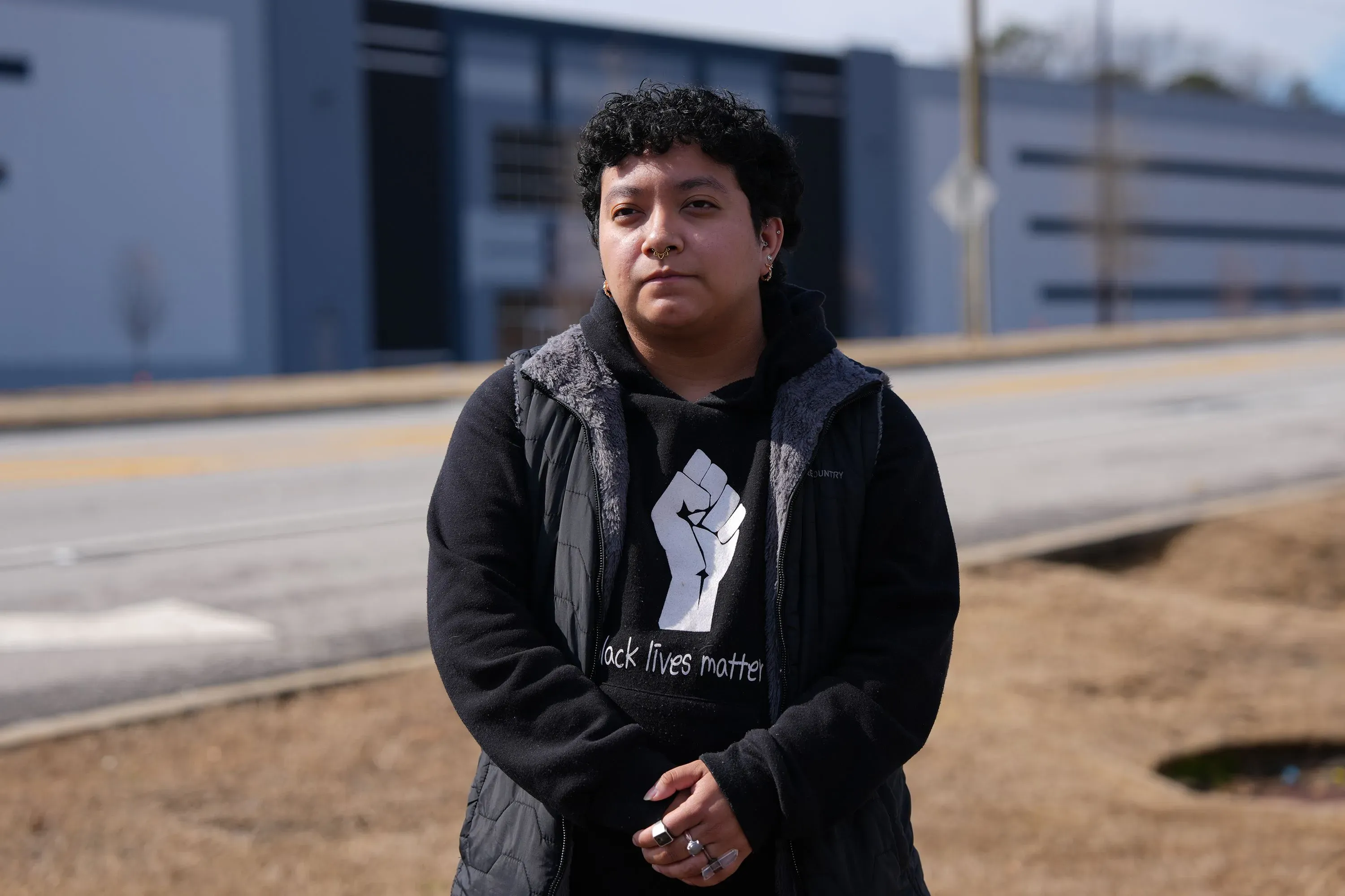 امرأة شابة ترتدي سترة داكنة وتظهر تعبيرًا جادًا، تقف أمام مستودع كبير، مع وجود لافتة "Black Lives Matter" على ملابسها، تعكس القلق بشأن مركز احتجاز المهاجرين القريب.