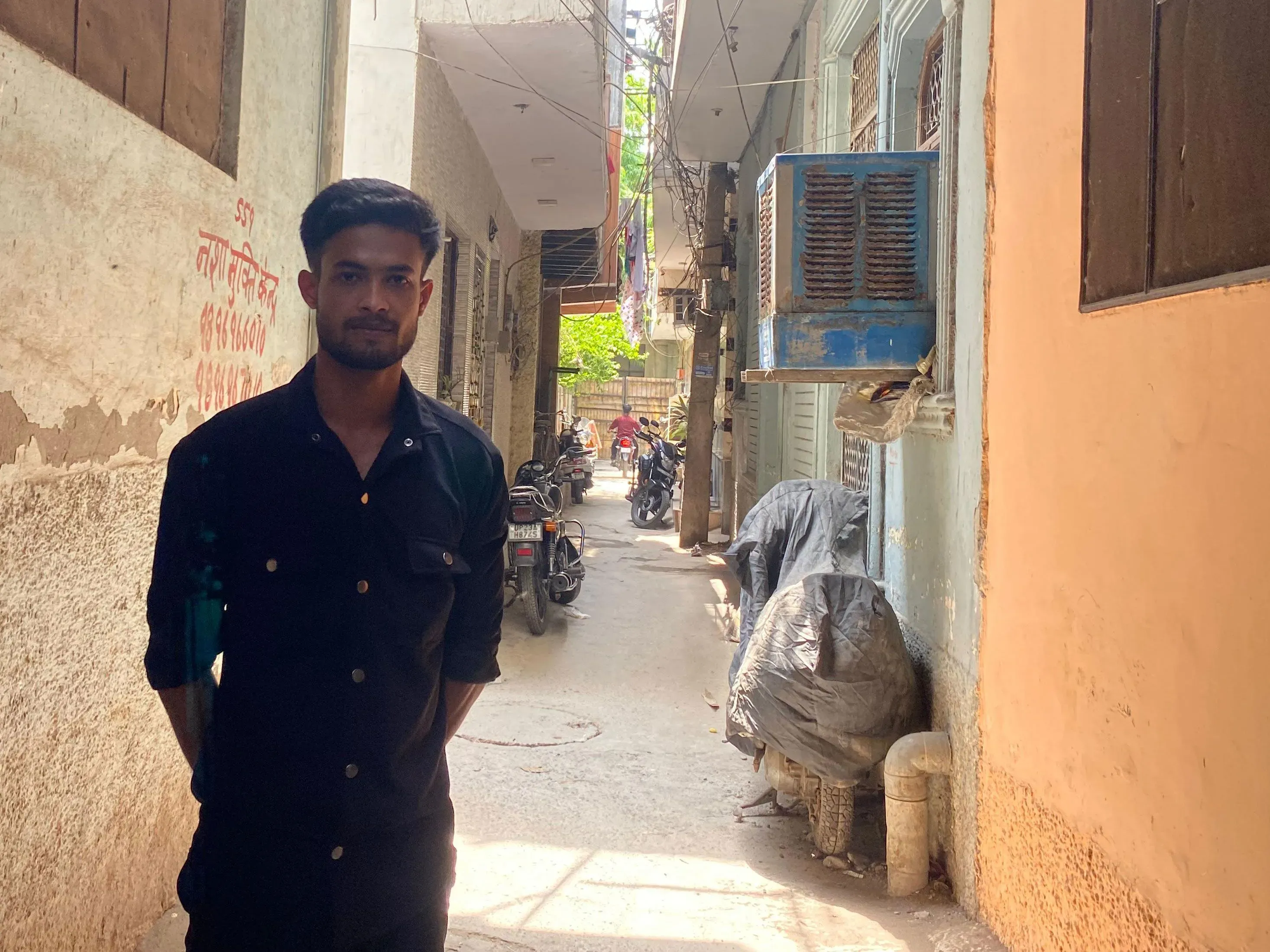 شاب يقف في زقاق ضيق في حي شاهين باغ، محاط بمبانٍ قديمة وأثاث مهمل، يعكس حياة اللاجئين الروهينغا في نيودلهي.