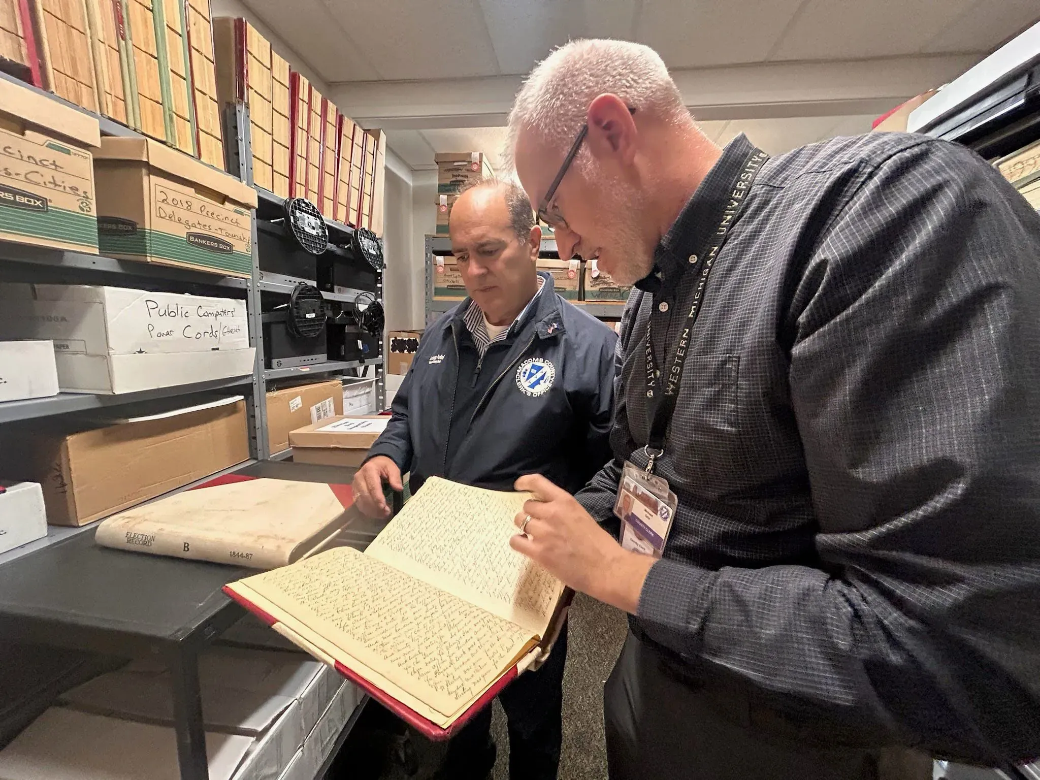 موظفان يتفقدان سجلات انتخابية في مكتب محلي، وسط صناديق تحتوي على مستندات متعلقة بالانتخابات، في سياق تدقيق الانتخابات.