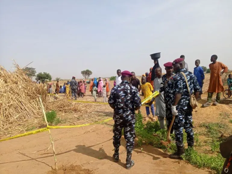 رجال الأمن يرتدون زيًا رسميًا، يراقبون موقعًا في منطقة ريفية، بينما يتجمع السكان المحليون في الخلفية، مما يعكس حالة من التوتر بعد الهجمات العسكرية.