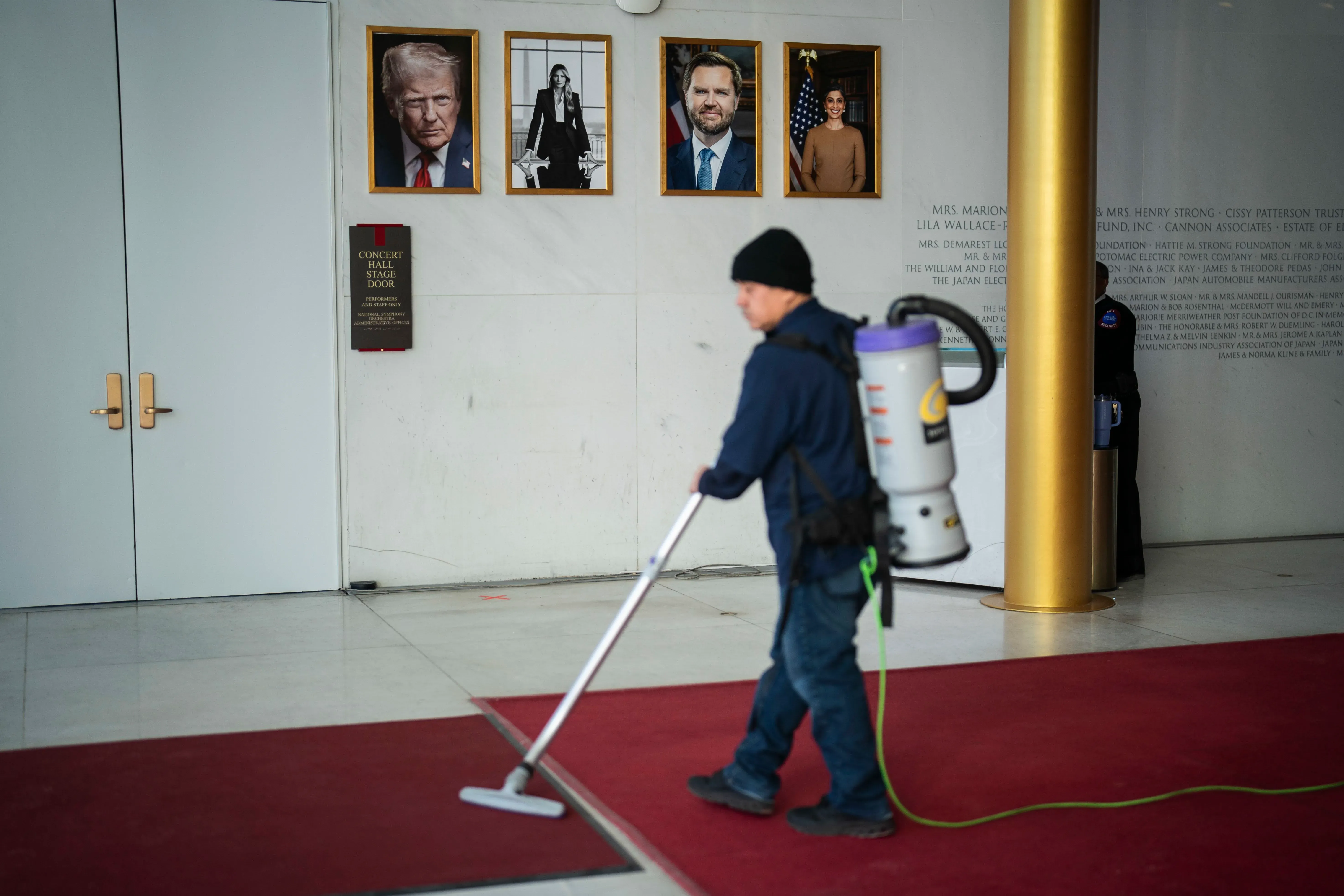 موظف يقوم بتنظيف السجادة في مركز كينيدي، مع وجود صور لترامب وشخصيات أخرى على الجدران، مما يعكس التغييرات الجارية في المؤسسة.
