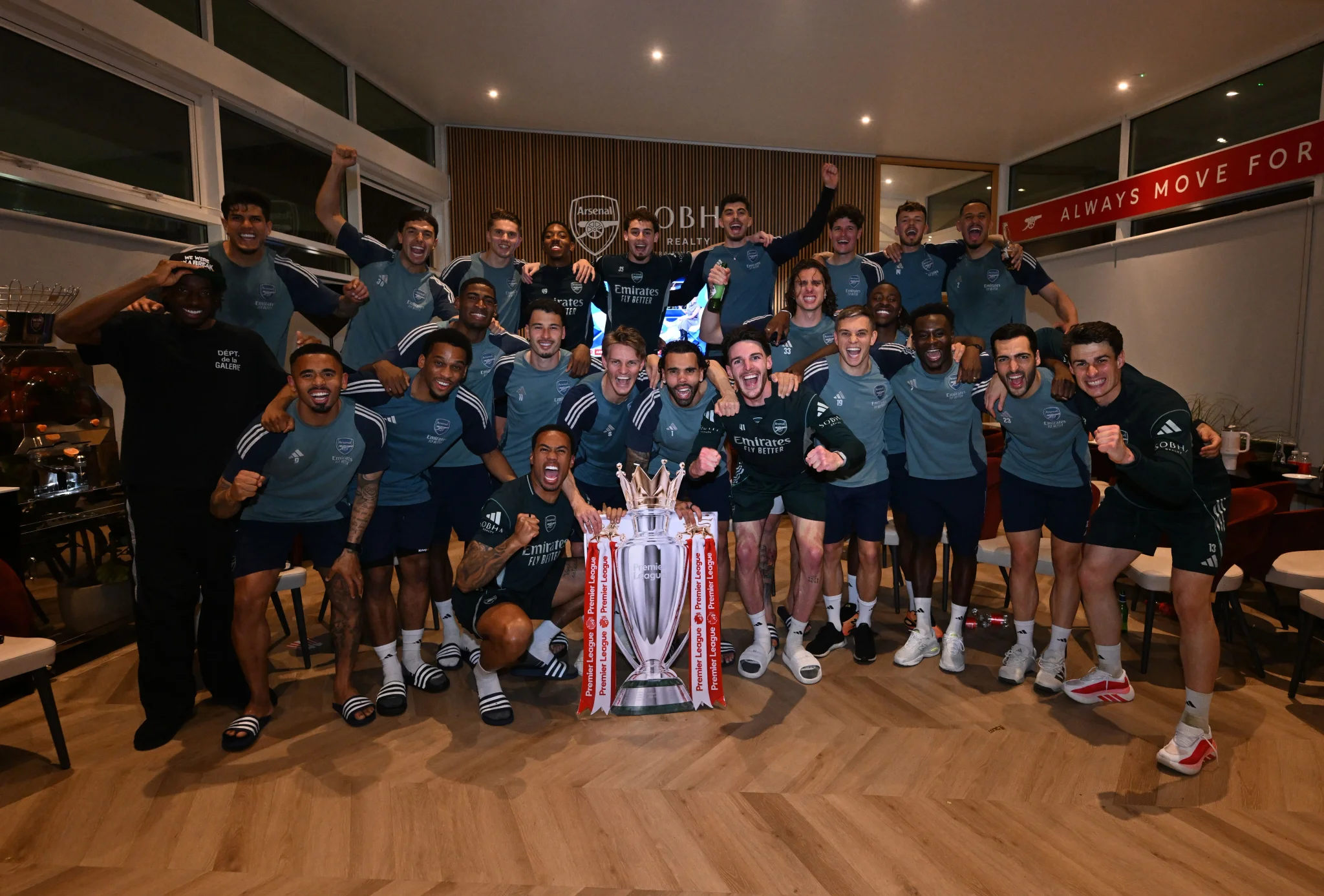 فريق Arsenal يحتفل بفوزه بلقب الدوري الإنجليزي الممتاز، محاطًا بكأس البطولة، مع تعبيرات الفرح والنشوة في أجواء احتفالية.