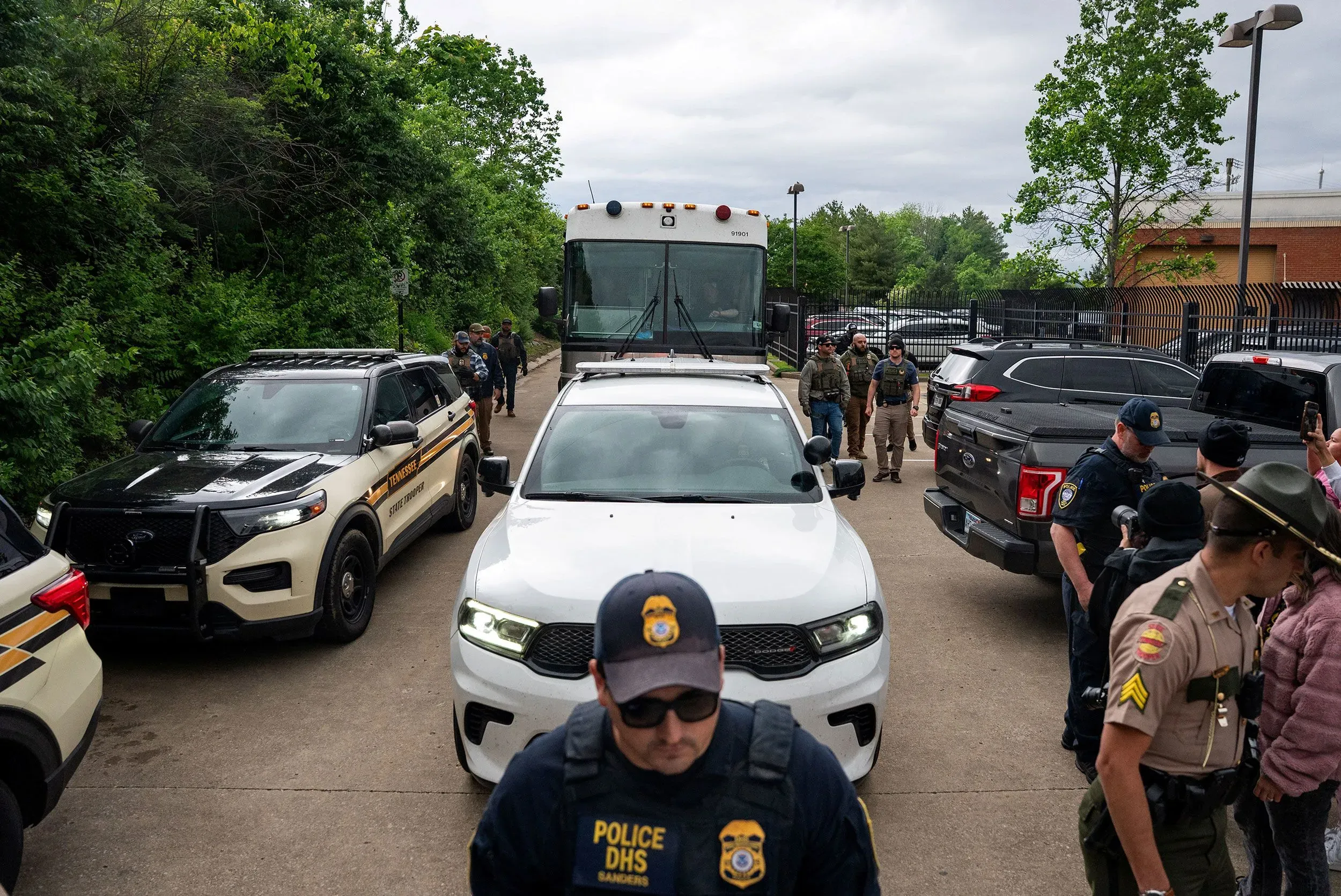مشهد يظهر مجموعة من ضباط إنفاذ القانون ومركباتهم في مكان مفتوح، مع شاحنة كبيرة في الخلفية، مما يعكس الأنشطة المتعلقة بعمليات الترحيل.
