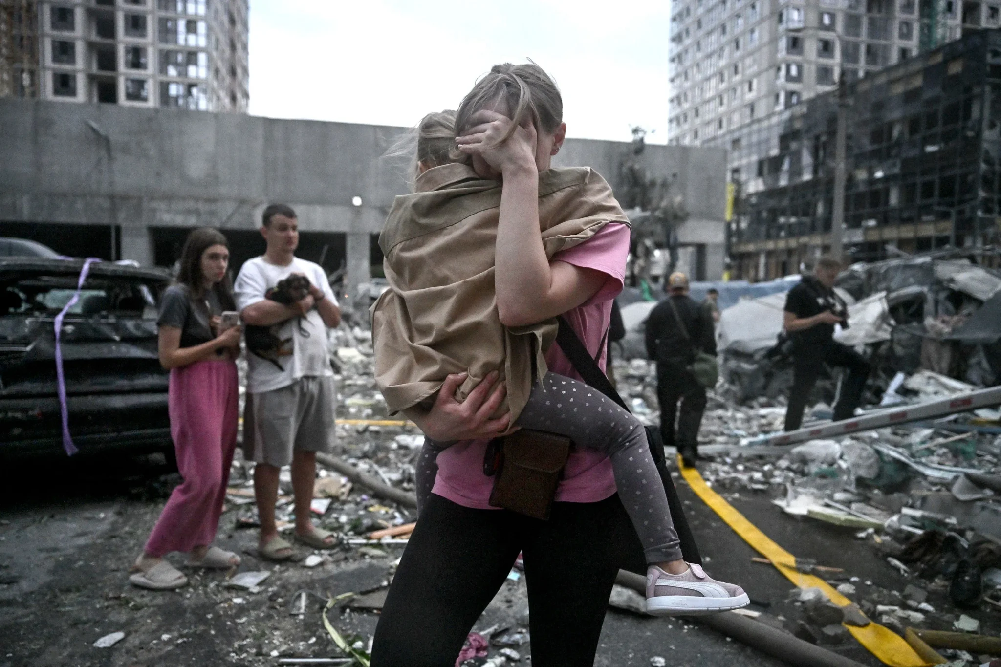 امرأة تحمل طفلة وسط أنقاض مبانٍ ومركبات مدمرة في مدينة أوكرانية بعد هجوم صاروخي روسي، مع ظهور مدنيين في الخلفية.