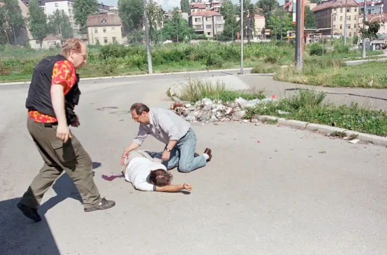 صورة تظهر شخصين يساعدان رجلًا ملقى على الأرض في منطقة حضرية، مع وجود حطام خلفهم، تعكس أجواء التوتر والعنف في سياق الأحداث التاريخية.