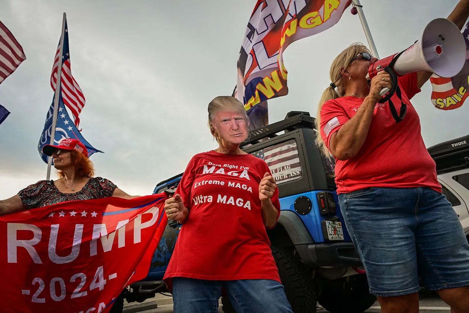 مؤيدون لترامب يرتدون قمصانًا تحمل شعاراته، يحملون لافتات، ويحتفلون في تجمع سياسي، مع وجود أعلام خلفهم.
