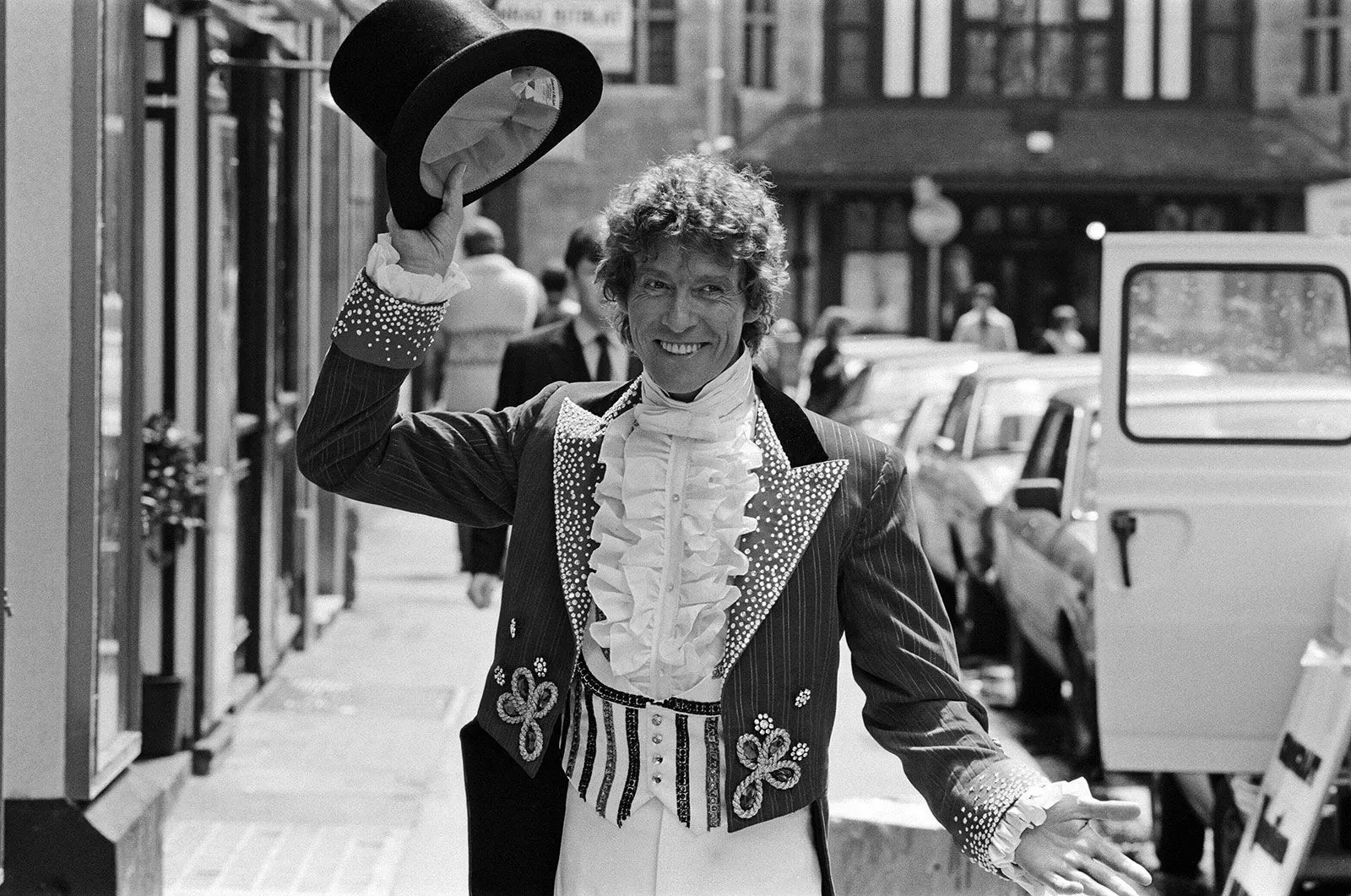 رجل يرتدي زيًا مميزًا مع قبعة، يبتسم ويرفع يده في تحية، بينما تظهر سيارات ومباني في الخلفية، مما يعكس أجواء احتفالية.