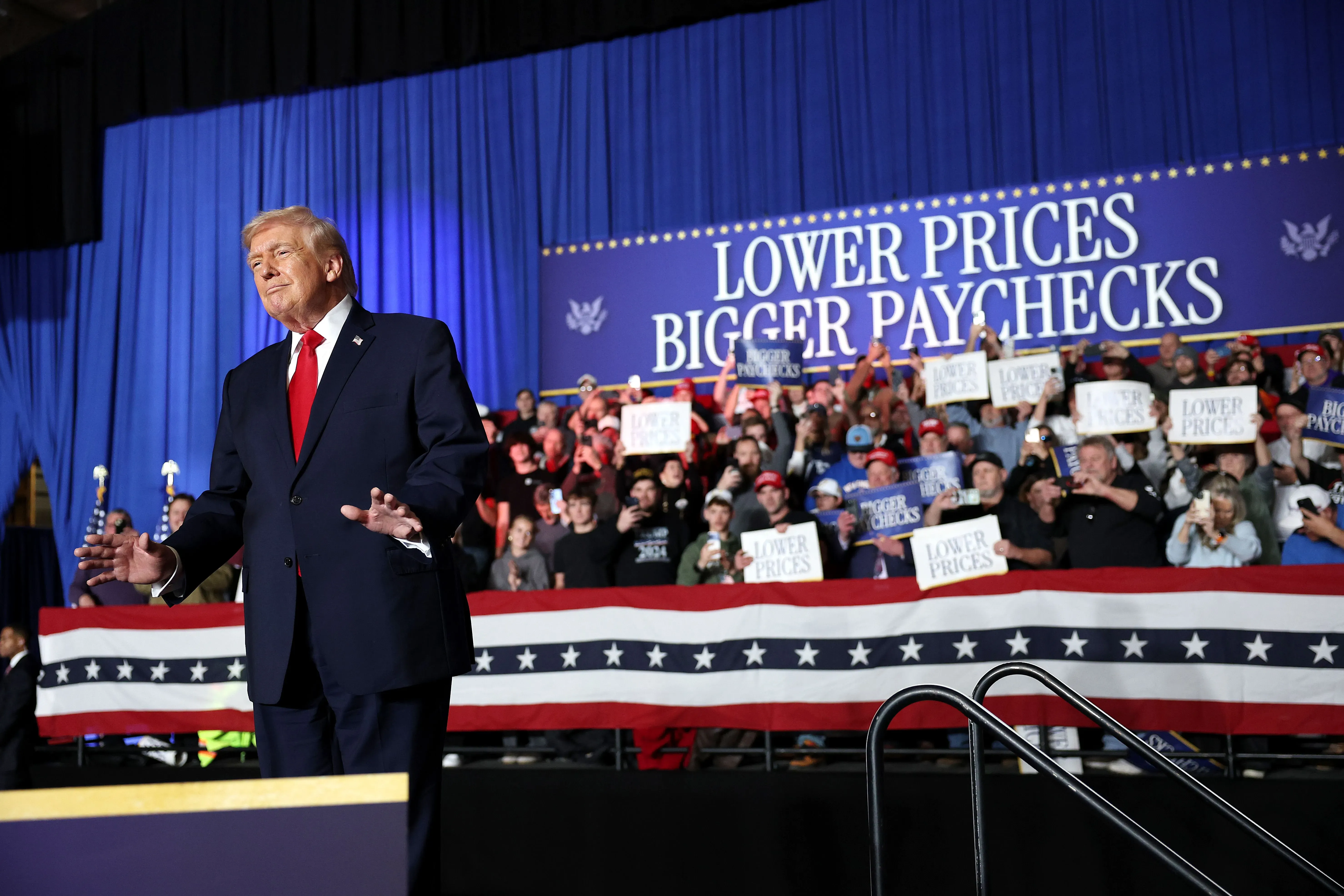 دونالد ترامب يتحدث أمام حشد من المؤيدين في تجمع انتخابي، مع لافتات تدعو إلى خفض الأسعار وزيادة الرواتب، مما يعكس قضايا سياسية حالية.