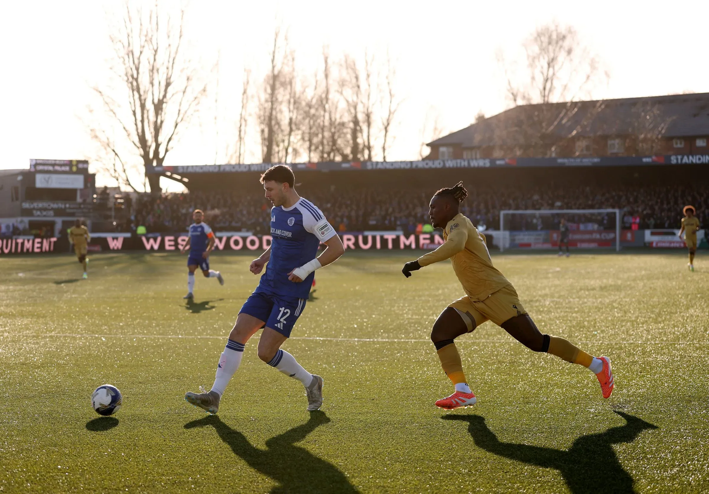 لاعبو ماكلسفيلد يتنافسون في مباراة كأس الاتحاد الإنجليزي، مع وجود مشجعين في المدرجات وأجواء مشمسة.