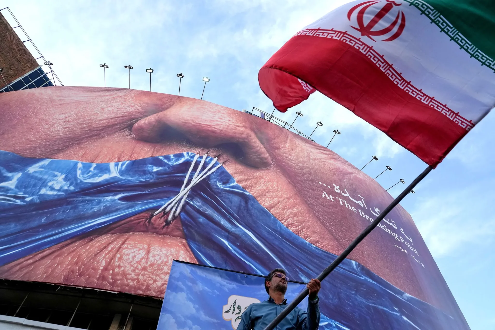 رجل يحمل علم إيران أمام لافتة ضخمة تُظهر وجه ترامب، تعبيراً عن التوترات بين الولايات المتحدة وإيران في سياق القمة المرتقبة في بكين.