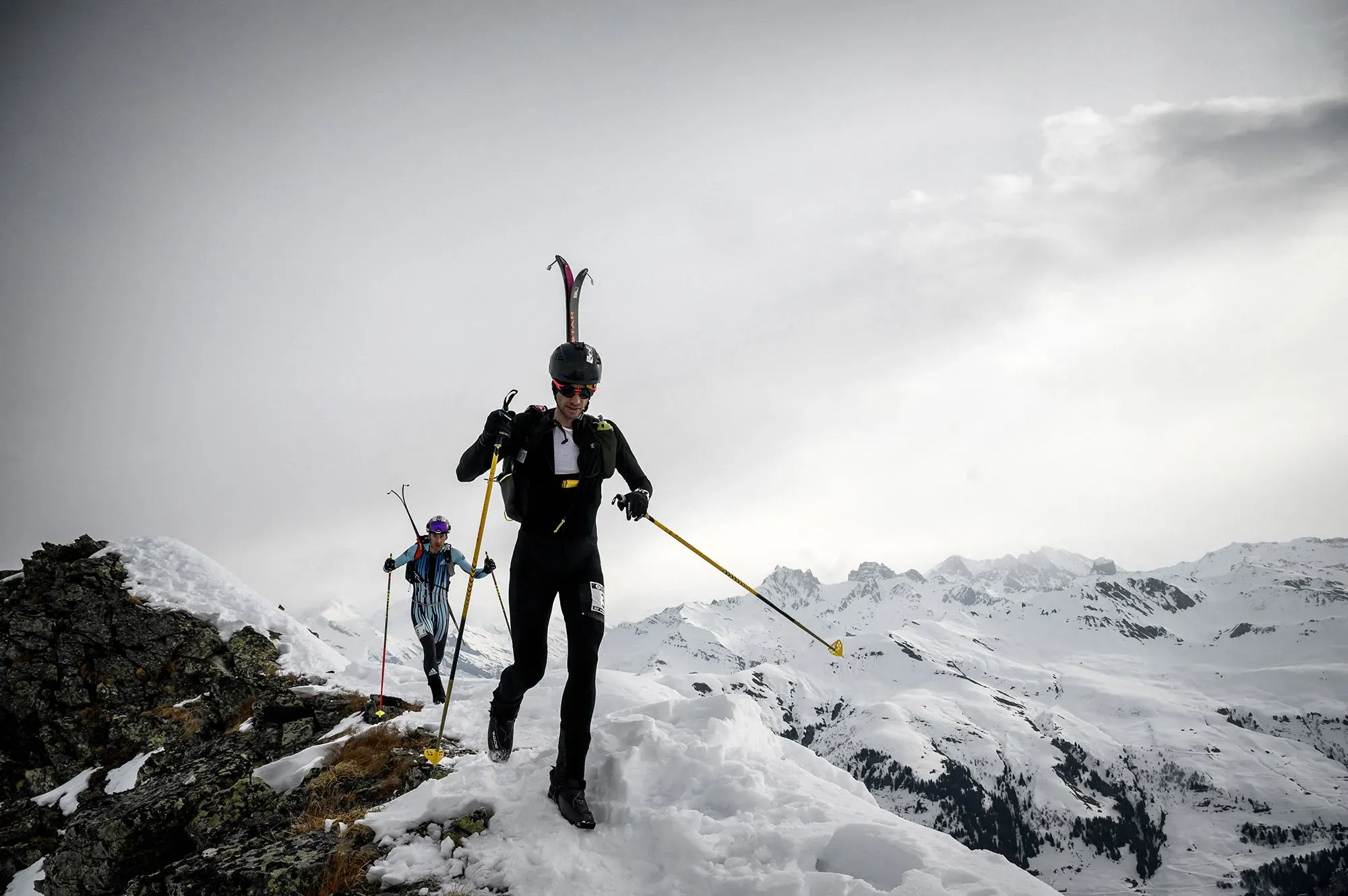 رجل يتسلق جبال الثلج الشاهقة، مرتديًا معدات التزلج والتسلق، مع خلفية من جبال مغطاة بالثلوج وسحب رمادية، يعكس تحديه للقدرة على التحمل والاستكشاف.