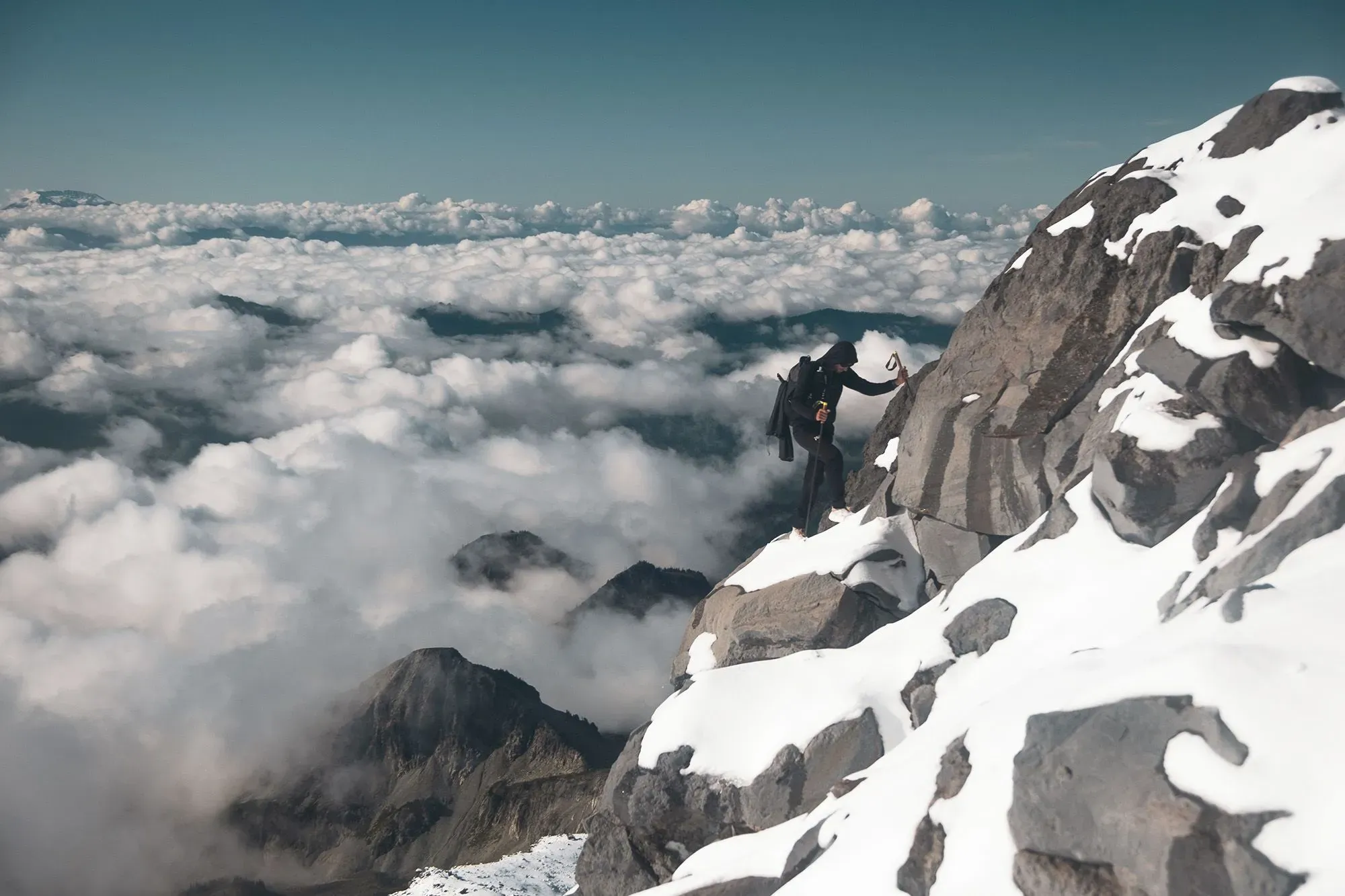 متسلق جبال يواجه قمة مغطاة بالثلوج والصخور، محاطاً بالغيوم والسماء الزرقاء، أثناء تسلقه الجبل في تحدٍ للقدرة على التحمل والاستكشاف.
