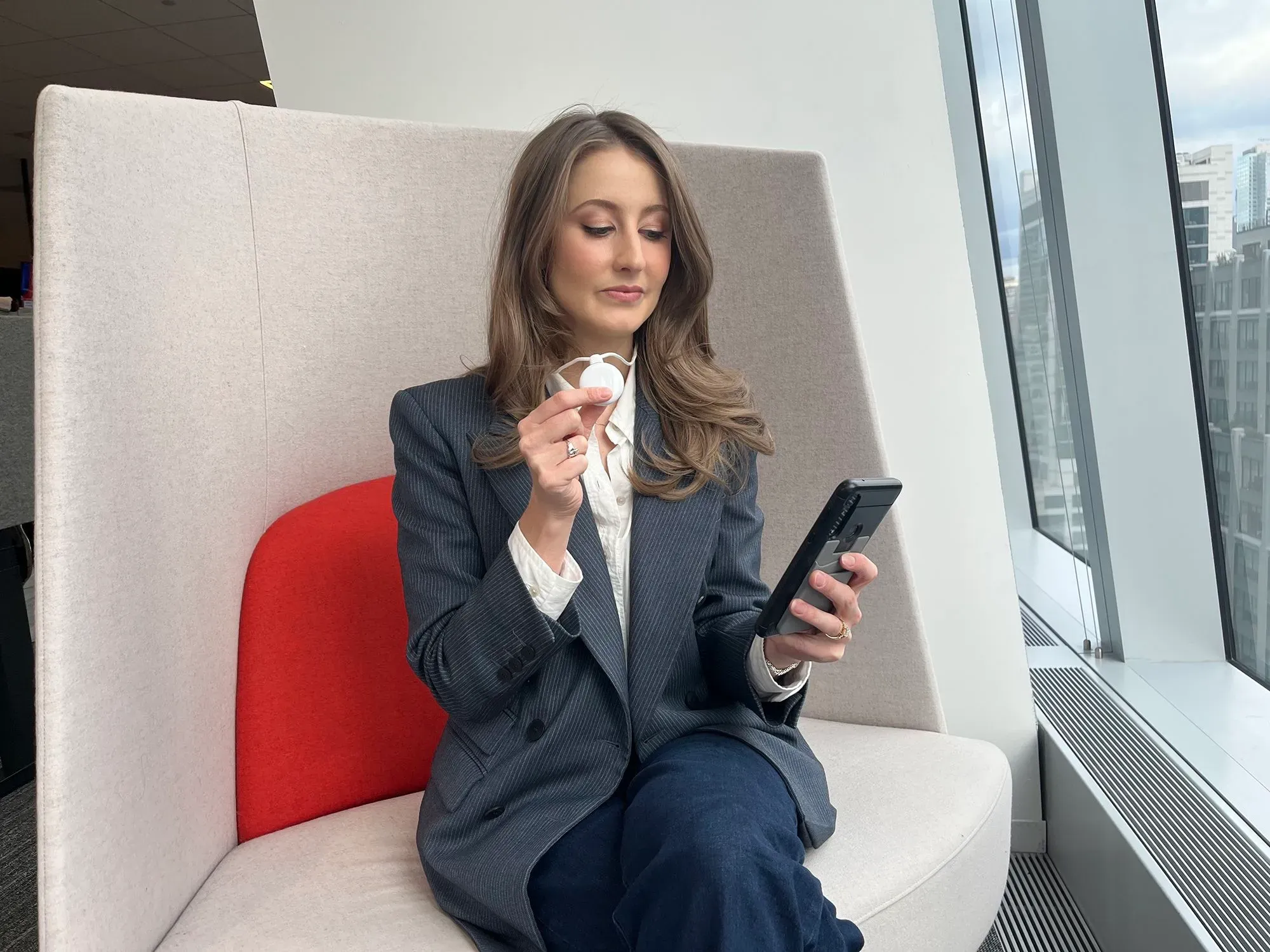 امرأة جالسة في منطقة مريحة، تحمل قلادة Friend الذكية بيدها وتستخدم هاتفها الذكي. تعكس الصورة ابتكار الذكاء الاصطناعي ودعمه للعلاقات الإنسانية.