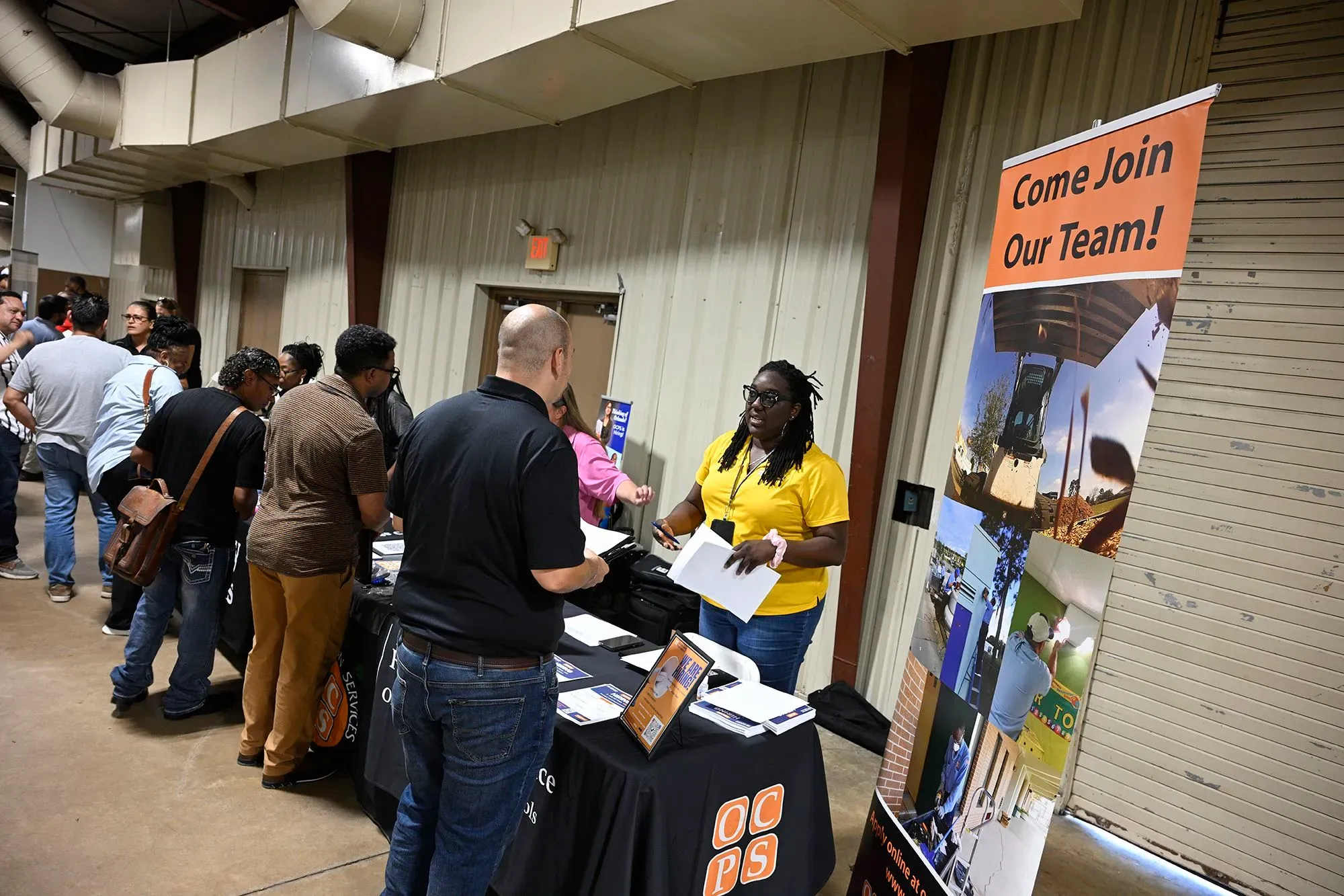 مشاركة في معرض توظيف، حيث يتجمع الباحثون عن عمل حول طاولة معلومات، مع وجود لافتة تدعوهم للانضمام إلى الفريق.