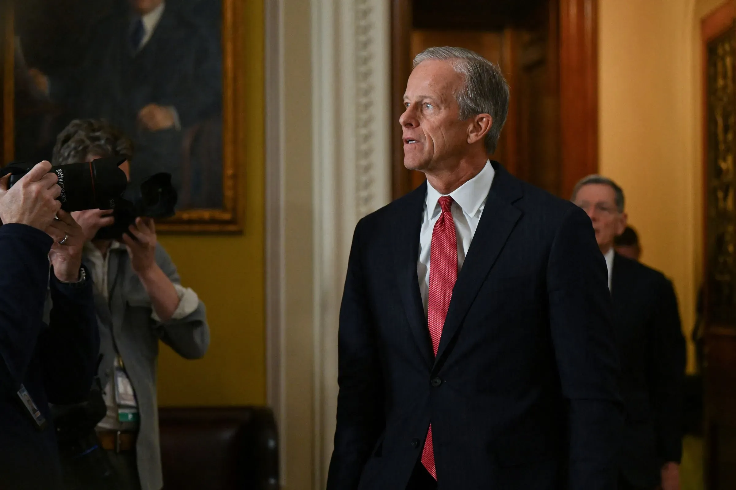 رجل يرتدي بدلة رسمية يسير في ممر برلماني، محاطًا بلوحات فنية، بينما يلتقط المصورون صورًا له.