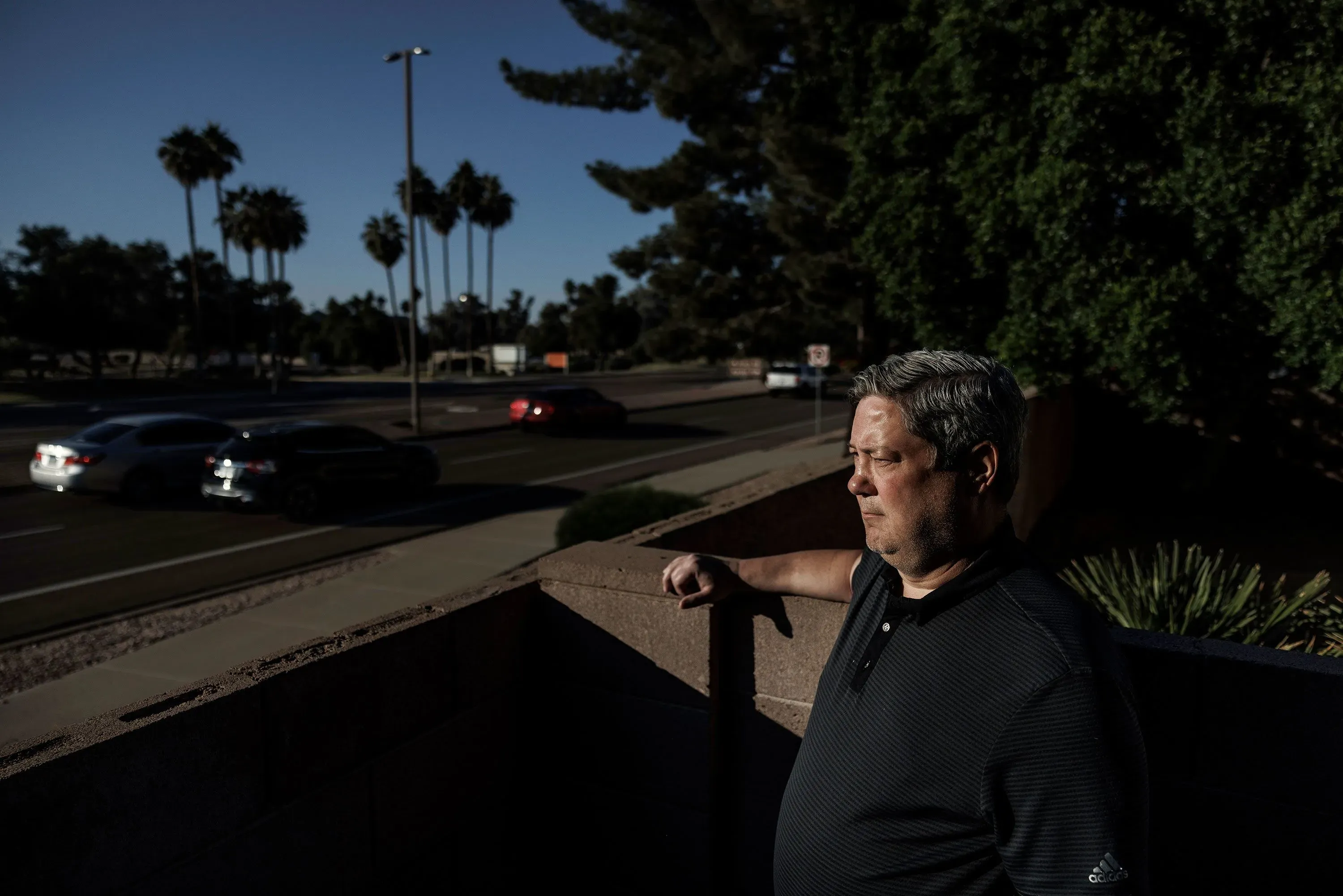 رجل يقف بجانب حافة جدار في مجمع سكني، يراقب الشارع المليء بأشجار النخيل والسيارات، مع تعبير جاد يعكس قلقه بشأن مراكز البيانات.