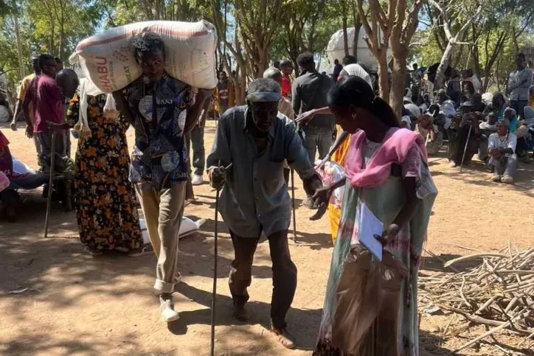 مشهد من قرية هيتساتس في تيغراي، حيث يساعد شاب مسنًا يحمل عكازًا، بينما ينتظر آخرون المساعدات الإنسانية وسط ظروف صعبة.