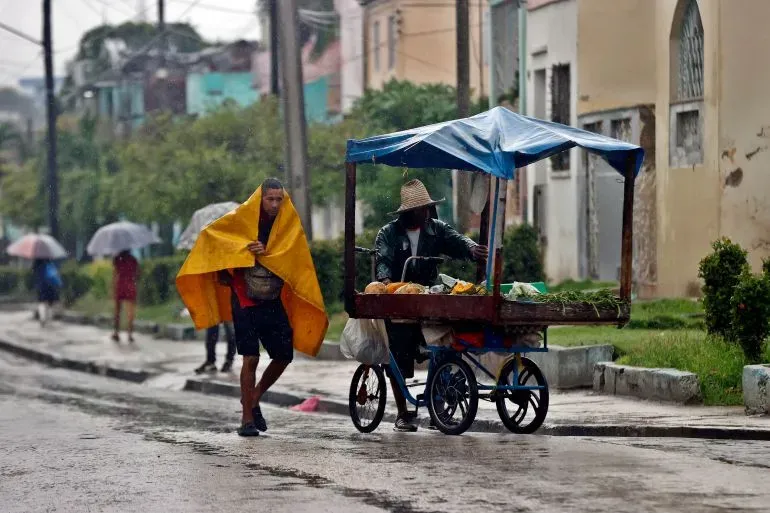 رجل يرتدي بطانية صفراء يسير في شارع ممطر بجوار بائع متجول يبيع الفواكه، وسط آثار إعصار ميليسا في كوبا.