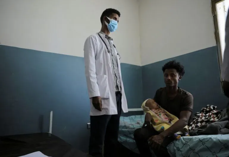 طبيب يرتدي معطفًا أبيض وقناعًا، يقف بجانب مريض يحمل طفلًا في غرفة طوارئ بمستشفى في أديس أبابا، مما يعكس تحديات القطاع الصحي في إثيوبيا.