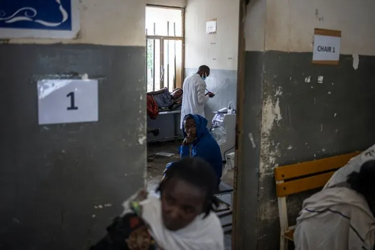 غرفة طوارئ في مستشفى بأديس أبابا، حيث يظهر طبيب يرتدي معطفًا أبيض، ومريض يجلس على الأرض، مع أثاث بسيط وظروف عمل صعبة.