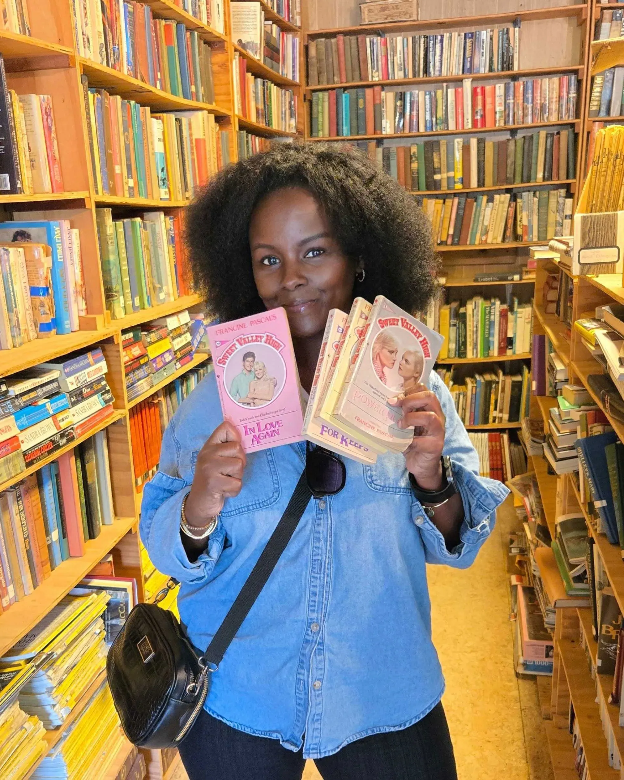 امرأة تحمل مجموعة من كتب "سويت فالي" في مكتبة صغيرة، محاطة برفوف مليئة بالكتب، تعكس شغفها بالقراءة واسترجاع ذكريات الطفولة.