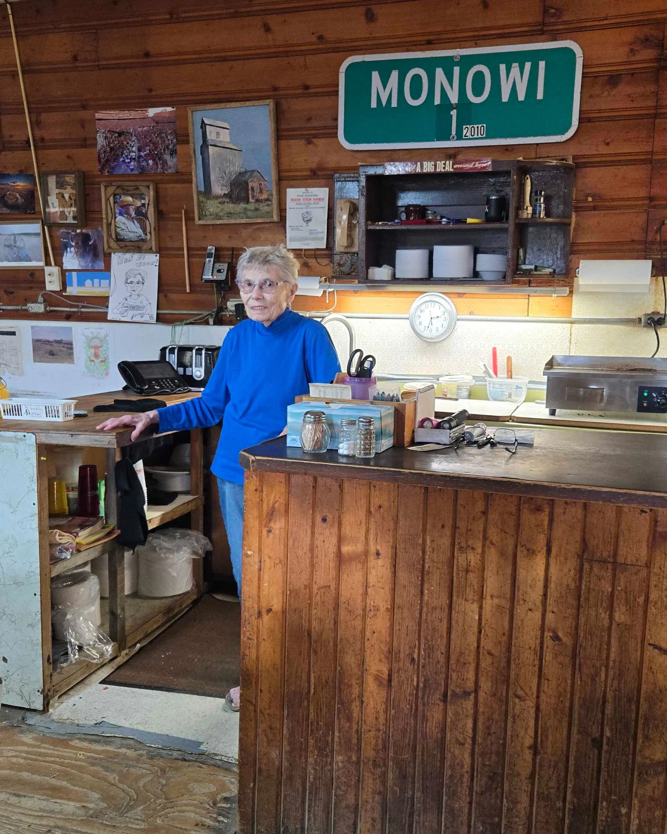 امرأة مسنّة ترتدي سترة زرقاء تقف خلف بار في حانة مونوي، مع لافتة تحمل اسم البلدة على الجدار، محاطة بأدوات المطبخ والكتب.