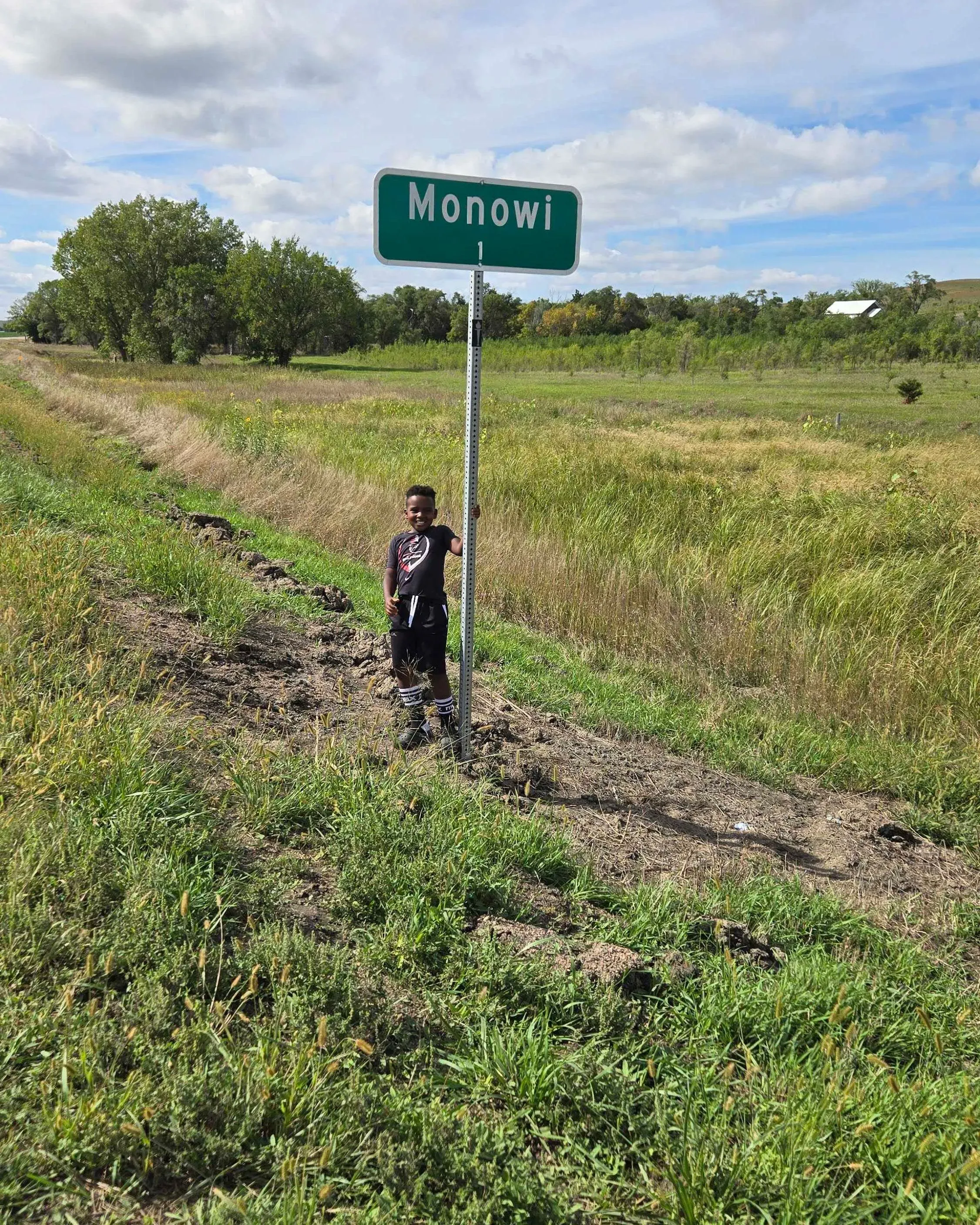طفل صغير يقف بجانب لافتة تشير إلى مدينة مونوي، مع منظر طبيعي من الحقول الخضراء والسماء الزرقاء، مما يعكس هدوء هذه المدينة الصغيرة.