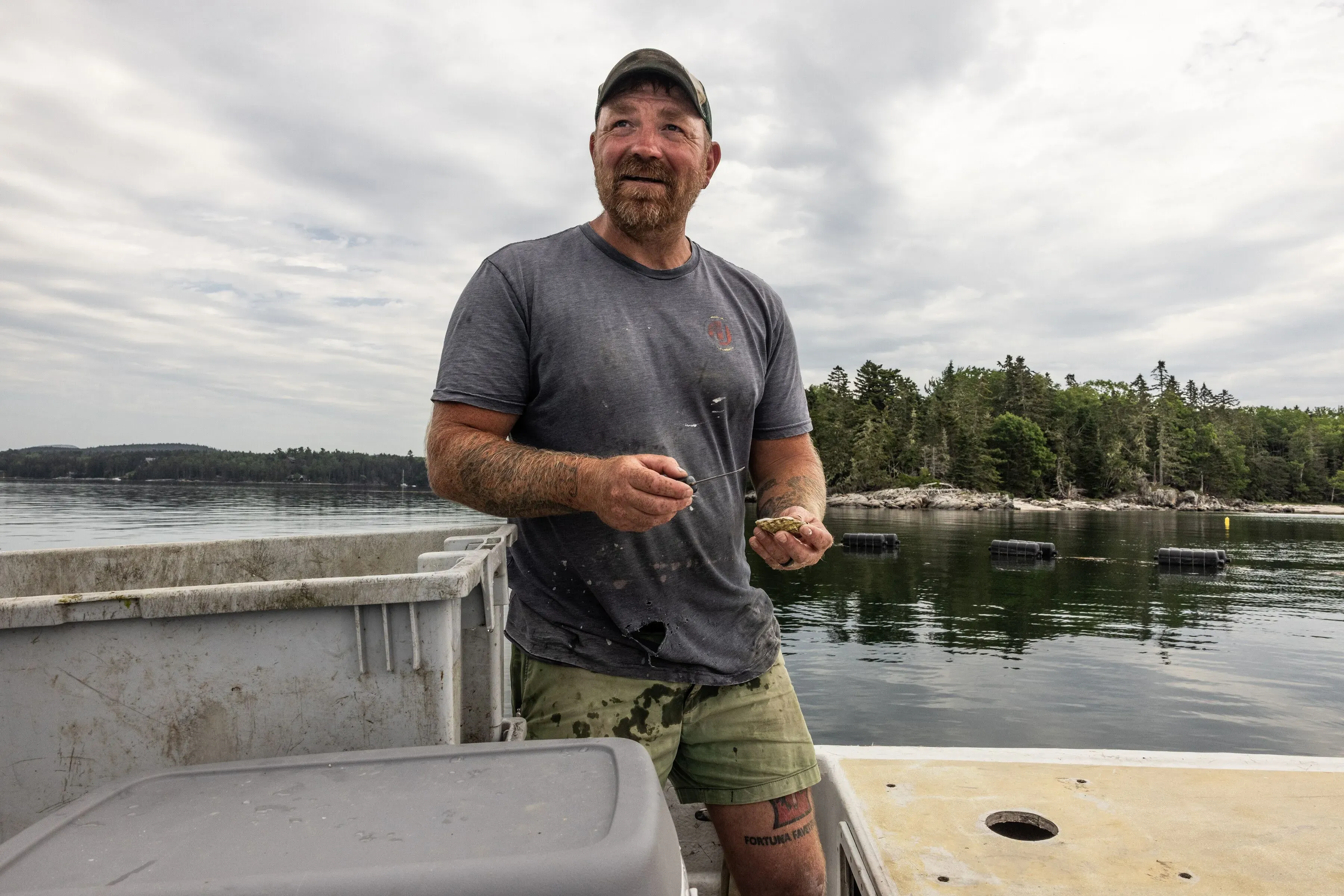 غراهام بلاتنر، مرشح ديمقراطي لمجلس الشيوخ في ولاية ماين، يقف في قارب مع المحار، محاطًا بمناظر طبيعية بحرية.