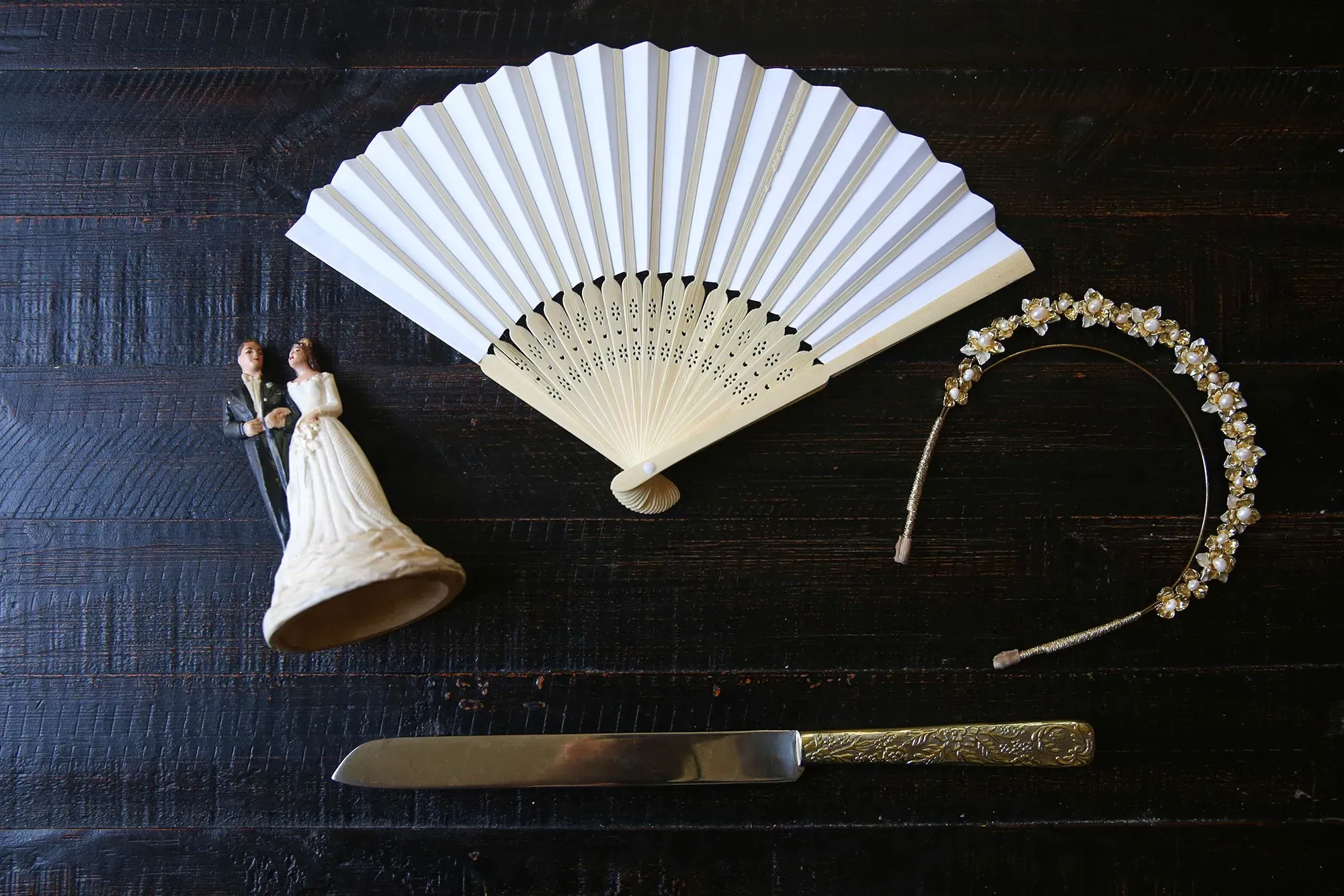 تتضمن الصورة مجموعة من عناصر حفلات الزفاف، تشمل تمثالين لعروسين، مروحة بيضاء، تاج مزين بالزهور، وسكين كعكة، مما يعكس أجواء الاحتفال.